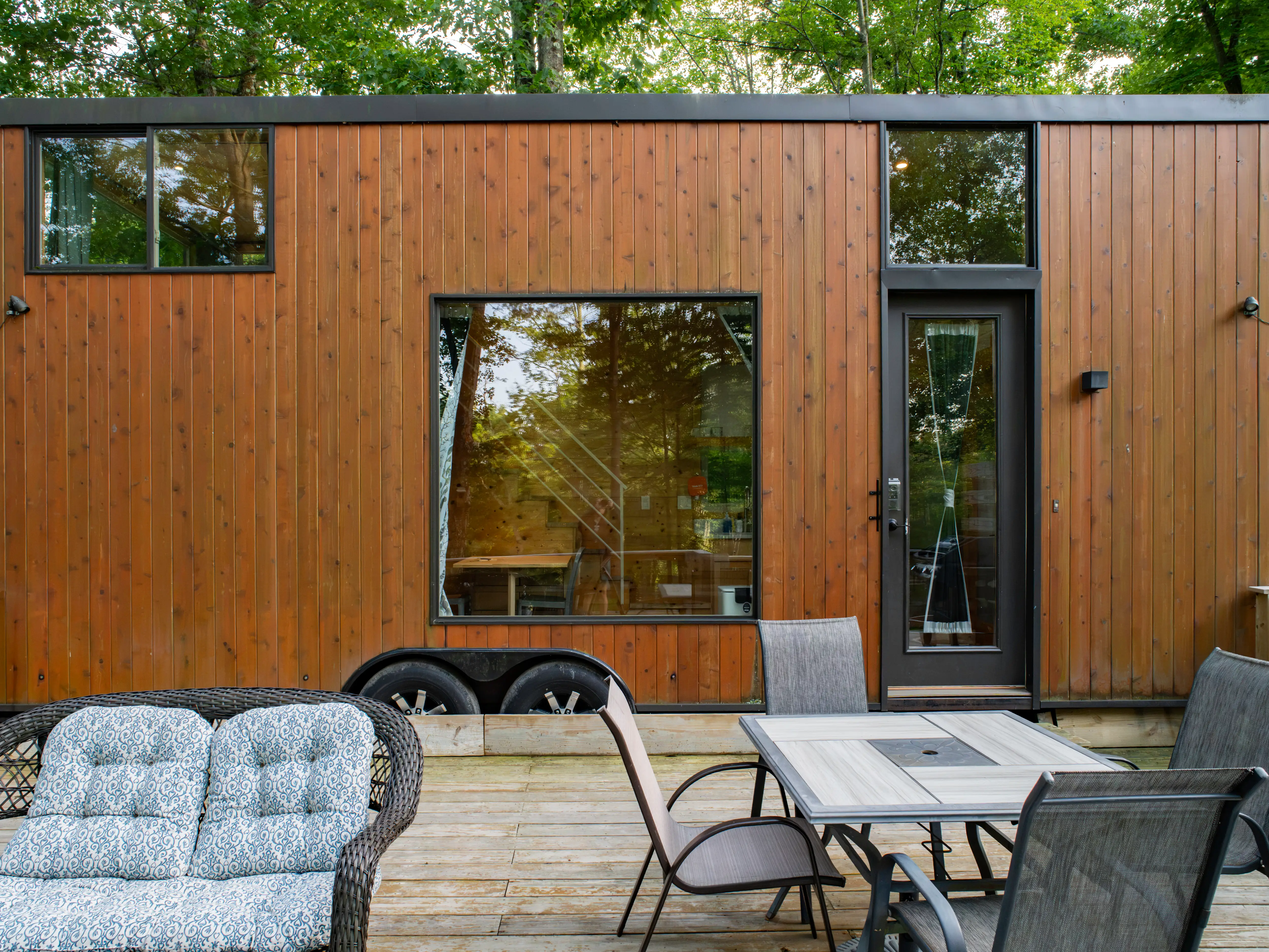 Wood-clad tiny house on wheels with large windows and patio furniture on a wooden deck.
