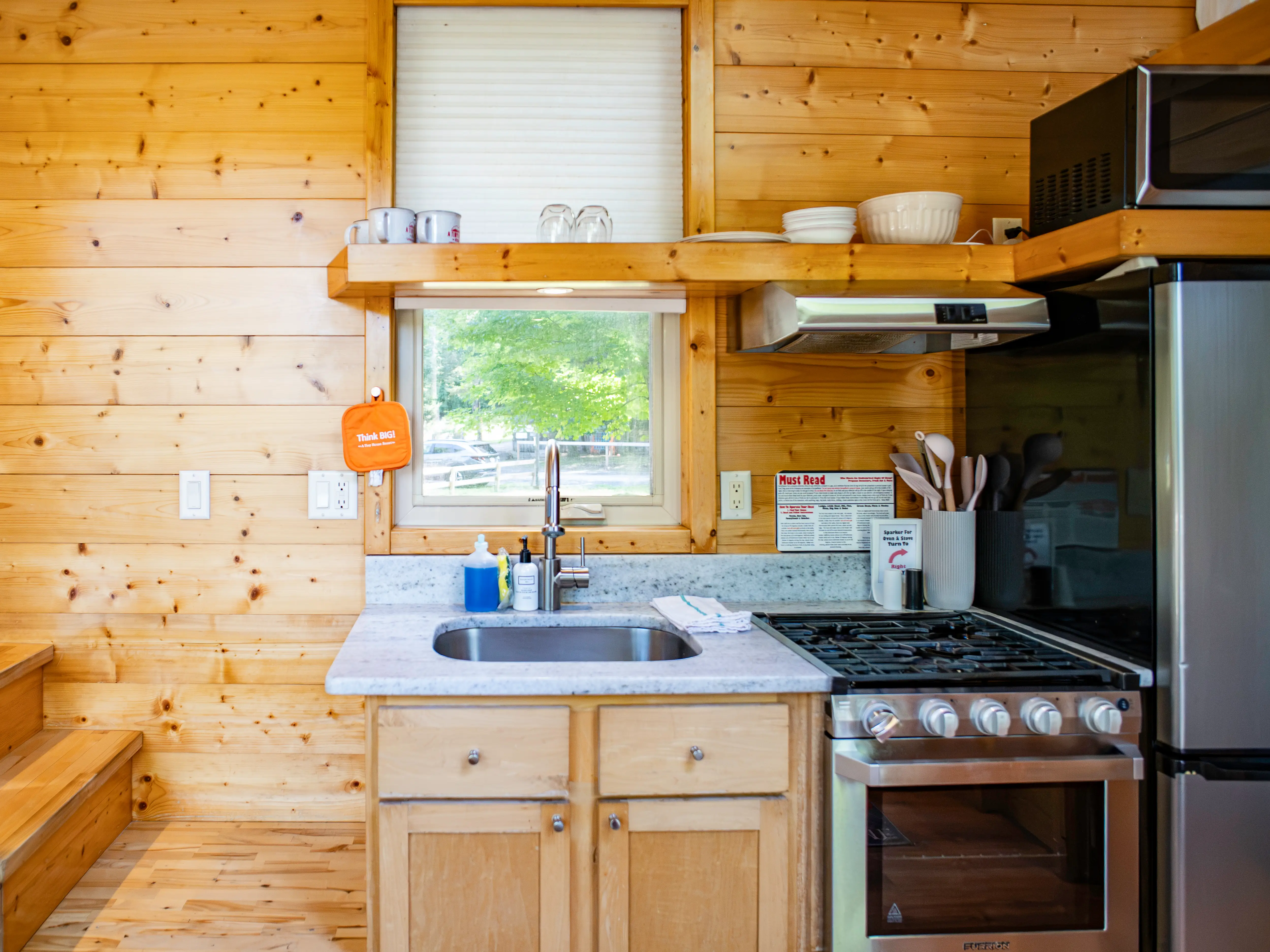 Compact wood-paneled kitchen with sink, gas stove, refrigerator, utensils, shelves, and a window above the counter.