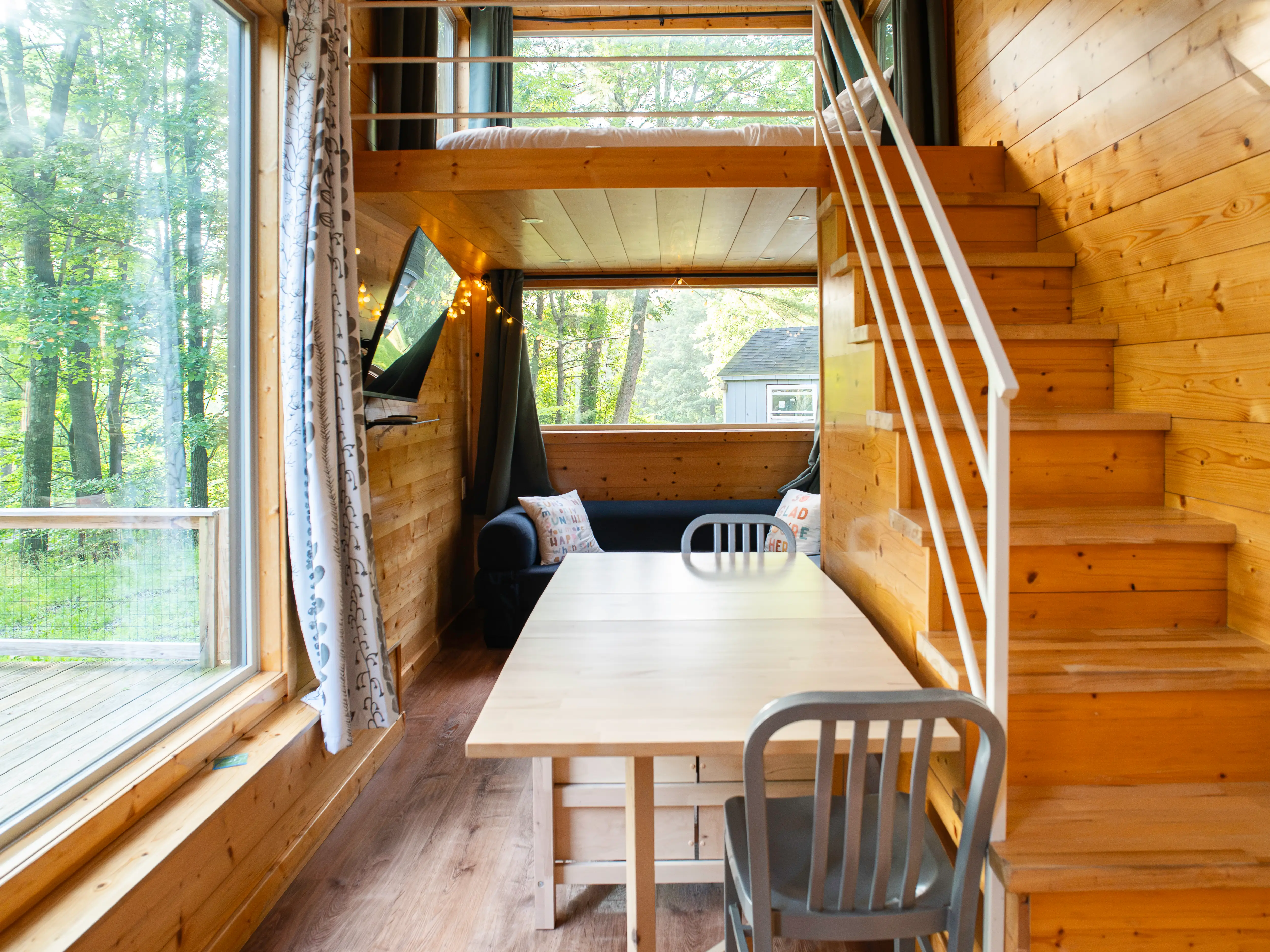 Wood-paneled tiny home interior with a loft staircase, dining table, chairs, sofa, and large window facing trees.