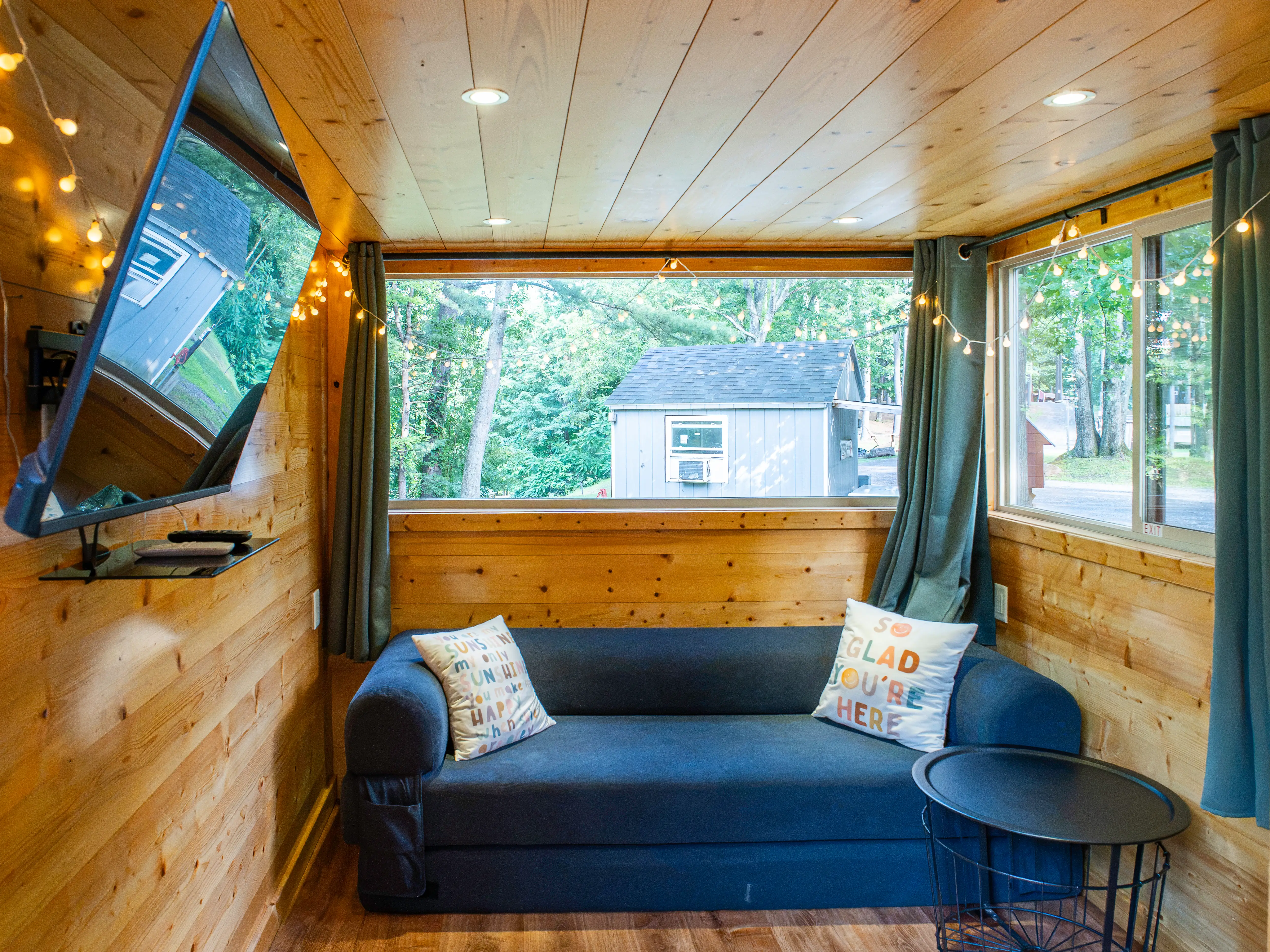 Wood-paneled tiny home living area with blue sofa, wall-mounted TV, large windows, curtains, string lights, and wooded views outside.