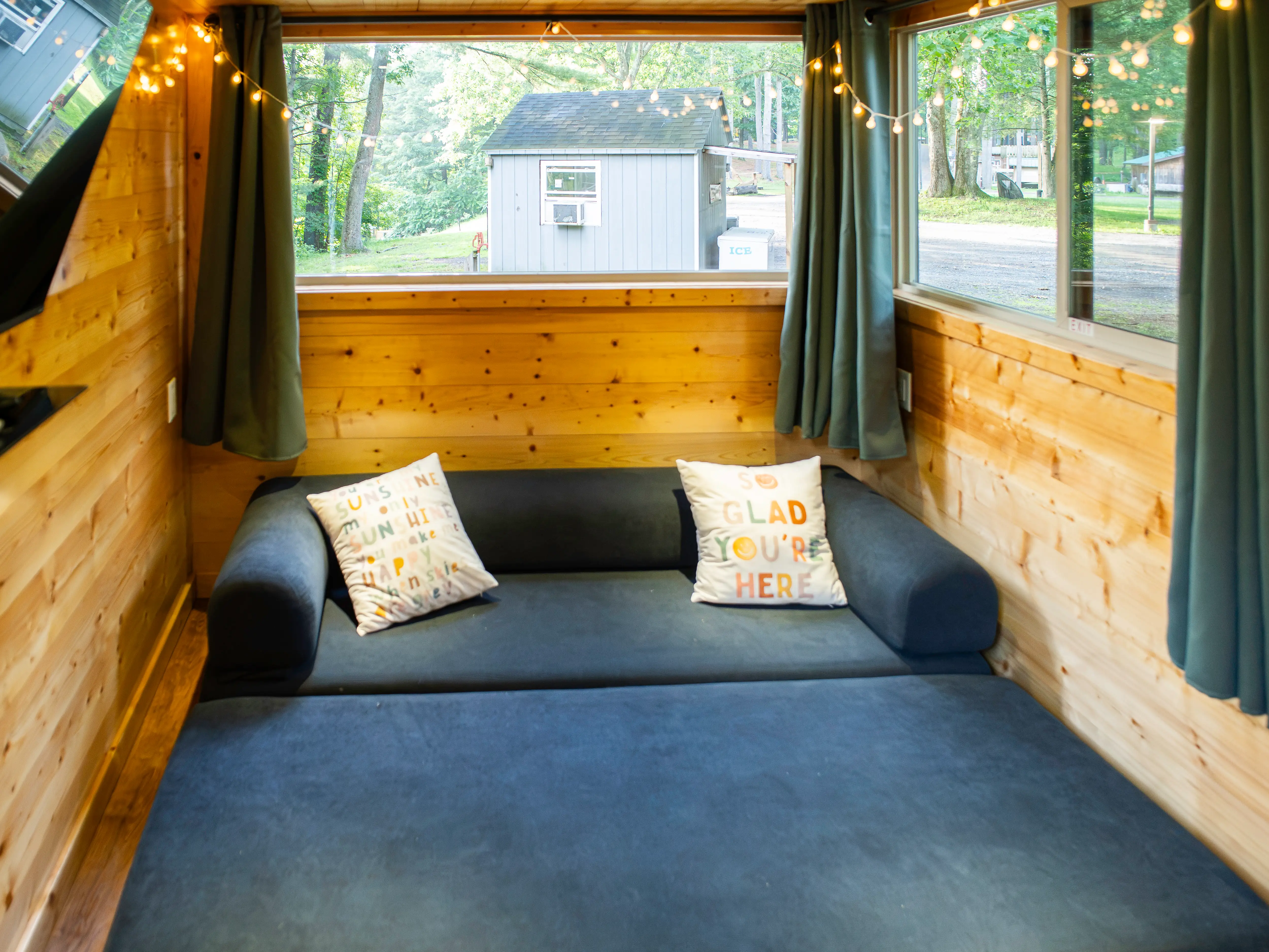 Wood-paneled tiny home room with a blue sofa bed, curtains, string lights, pillows, and large windows facing trees.