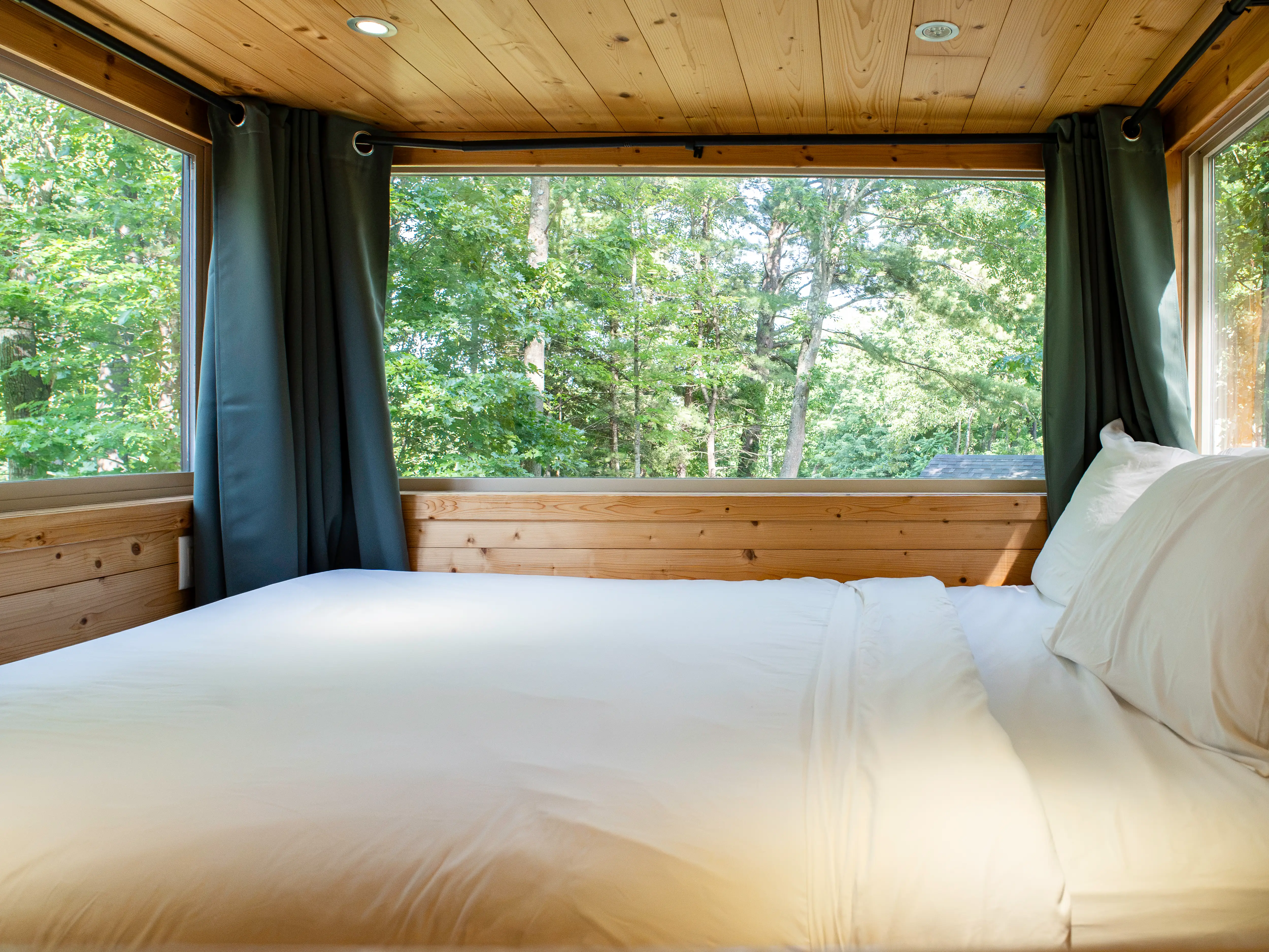 White bed inside a wood-paneled tiny home loft with large windows looking out onto green trees.