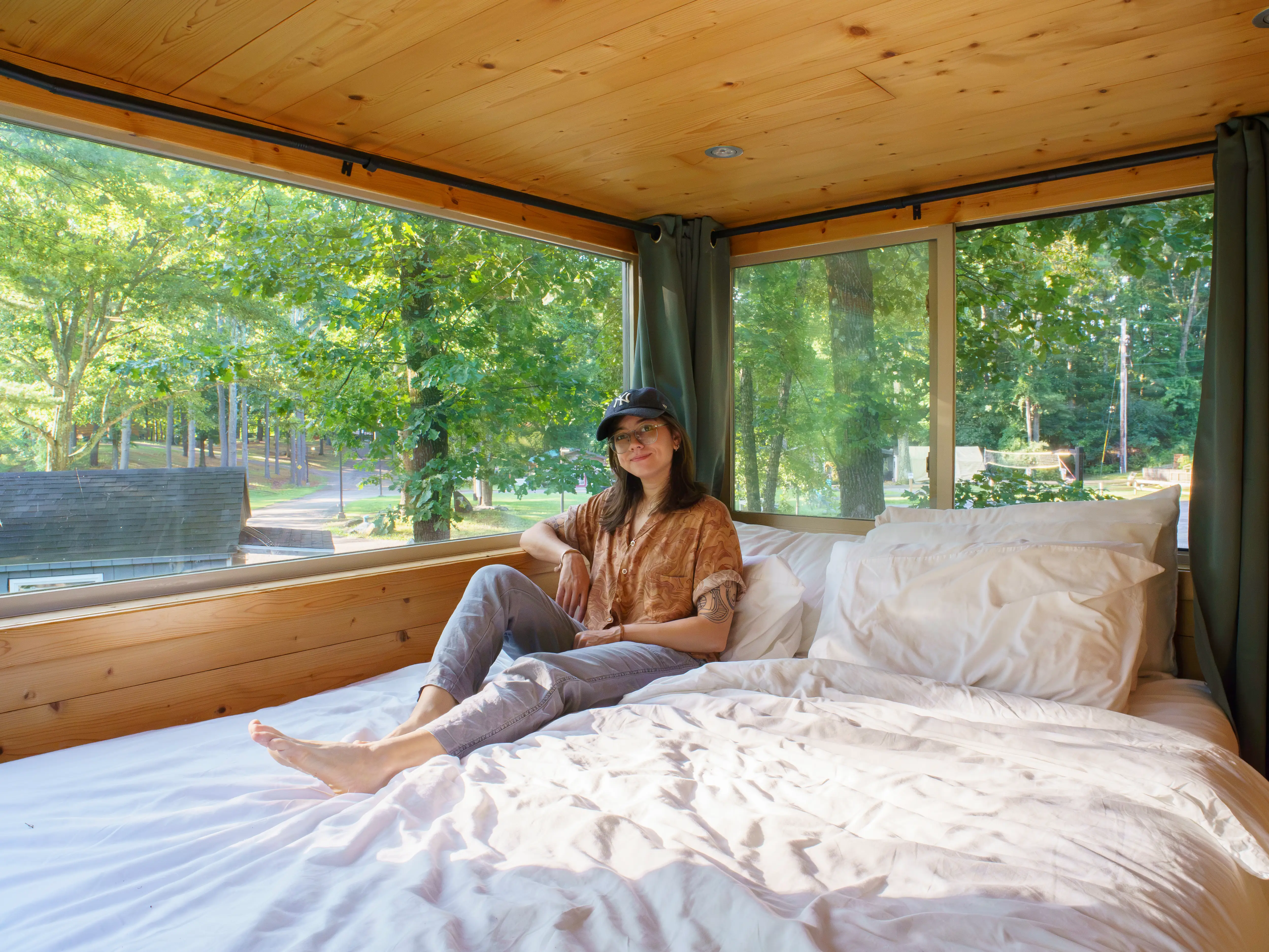 The author sits on a white bed inside a wood-paneled room with large windows overlooking green trees.