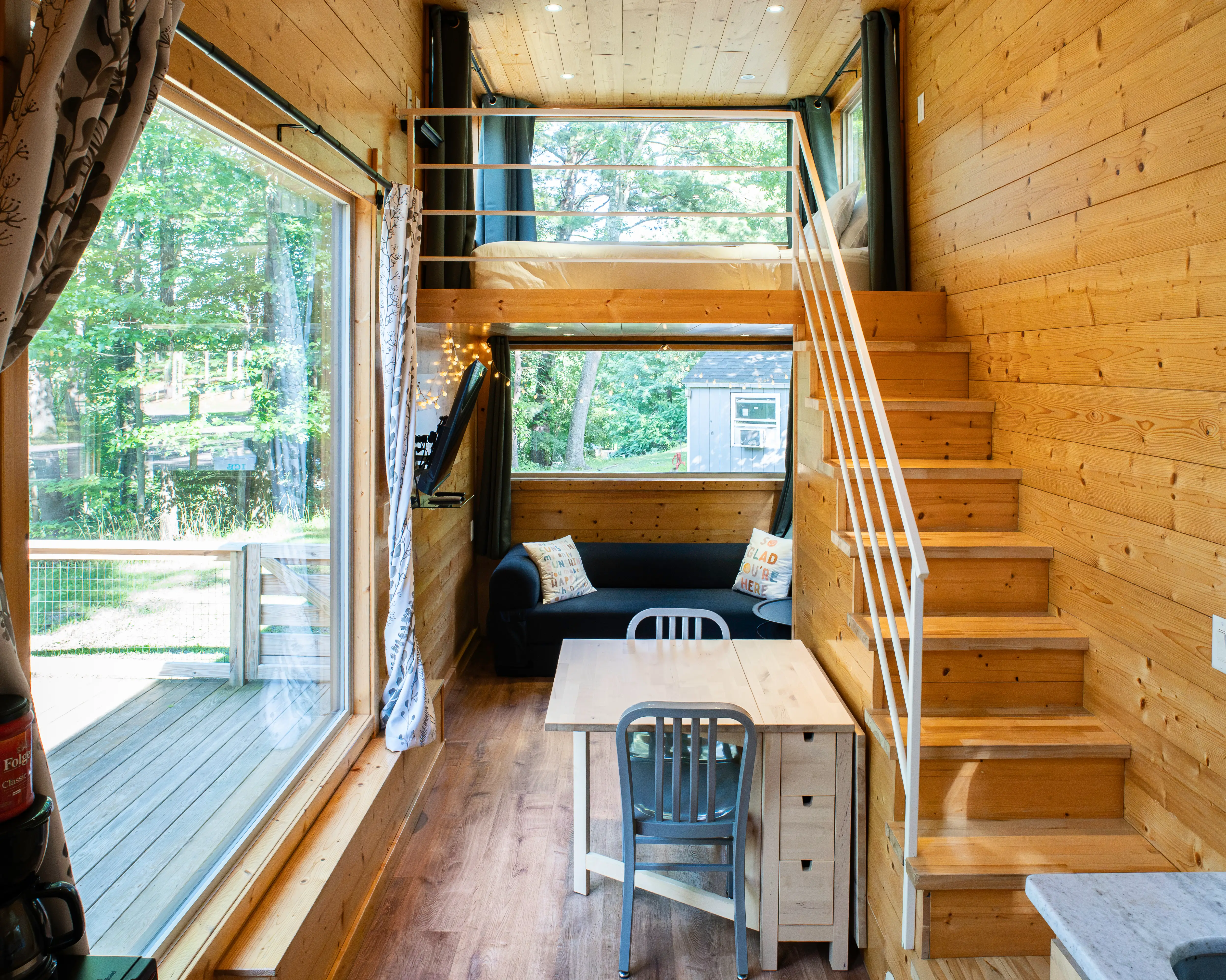 Compact wood-paneled tiny home interior with a loft bed, a staircase, a desk, a sofa, a large window, and a forest view.