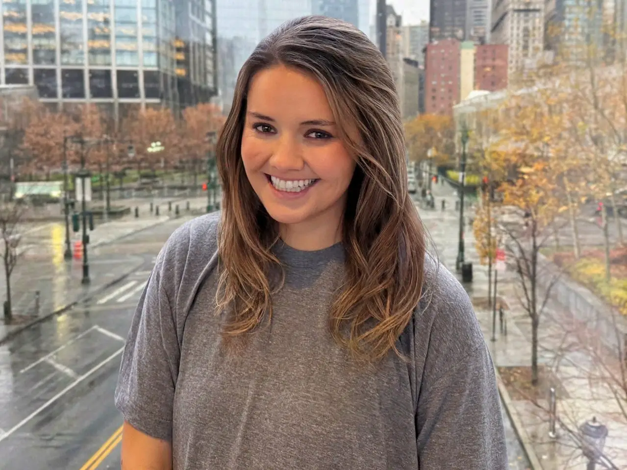 Smiling woman in a gray shirt poses indoors with a rainy city street and buildings visible behind them.