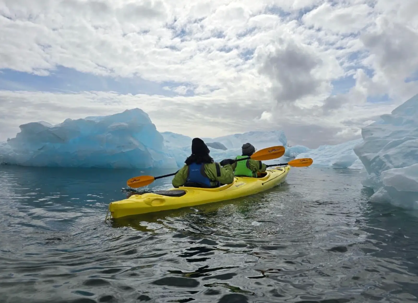 Two kayakers paddle a yellow tandem kayak through icy water between large blue-white icebergs.