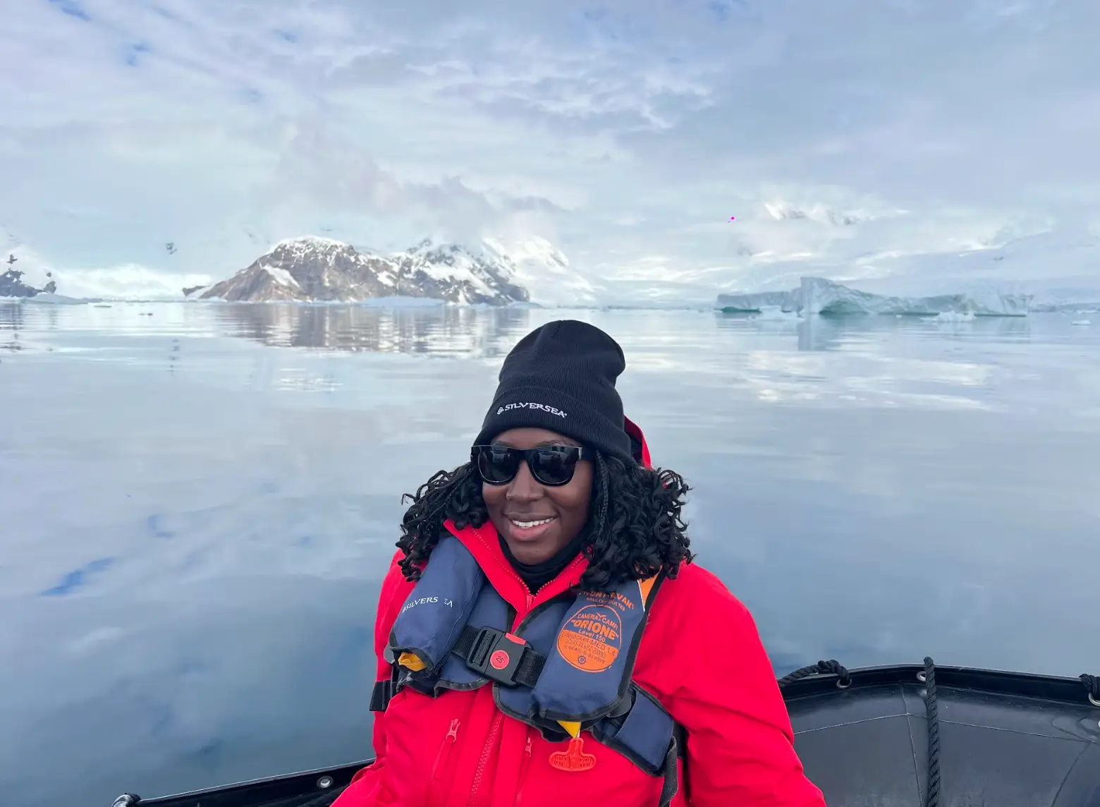 The writer wearing a life vest and a red jacket and sitting on a cruise in front of snowcapped mountains.
