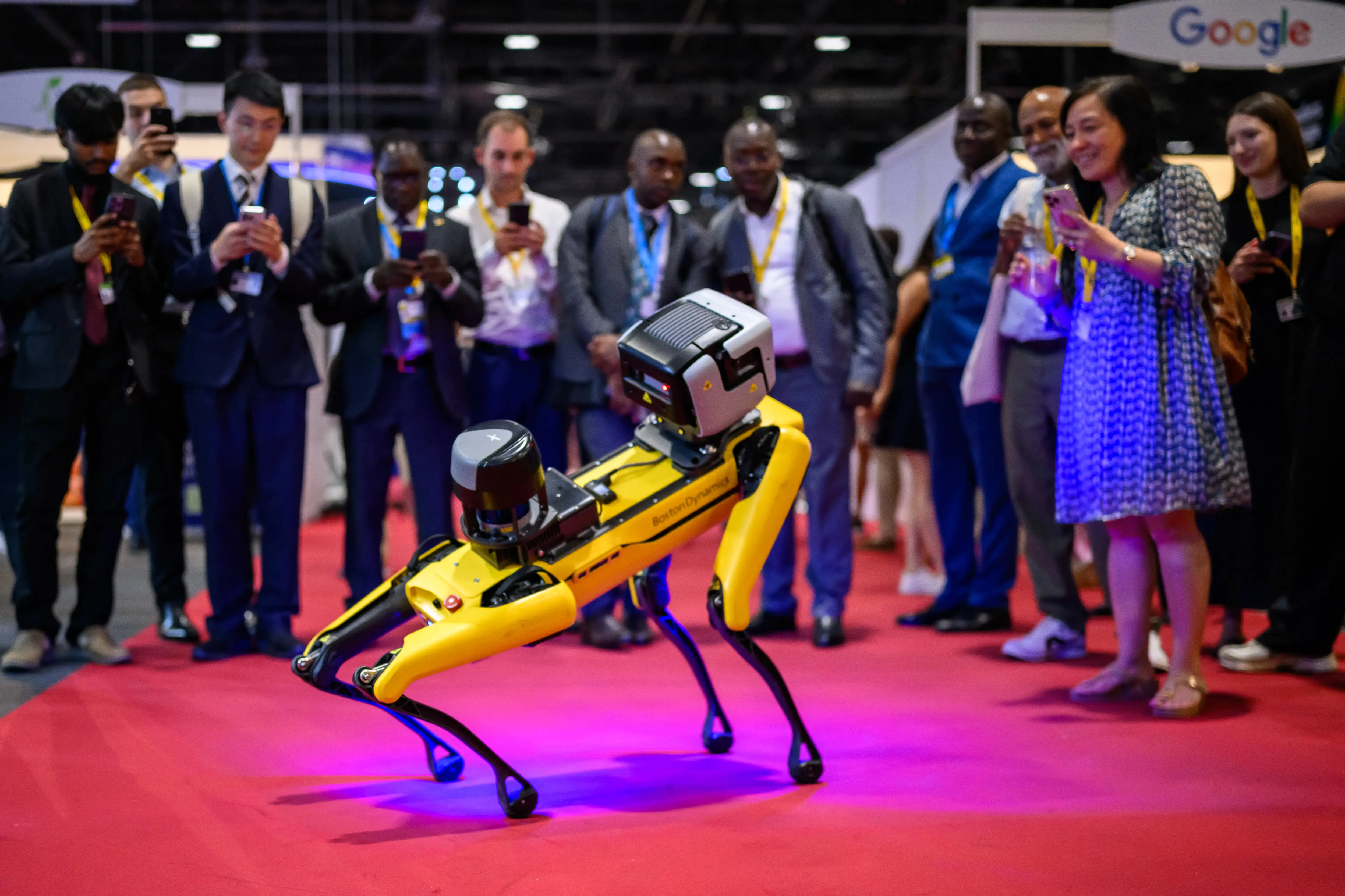 A yellow-and-black robotic dog created by Boston Dynamics stands on a red carpet with people standing around it and taking photos with smartphones.