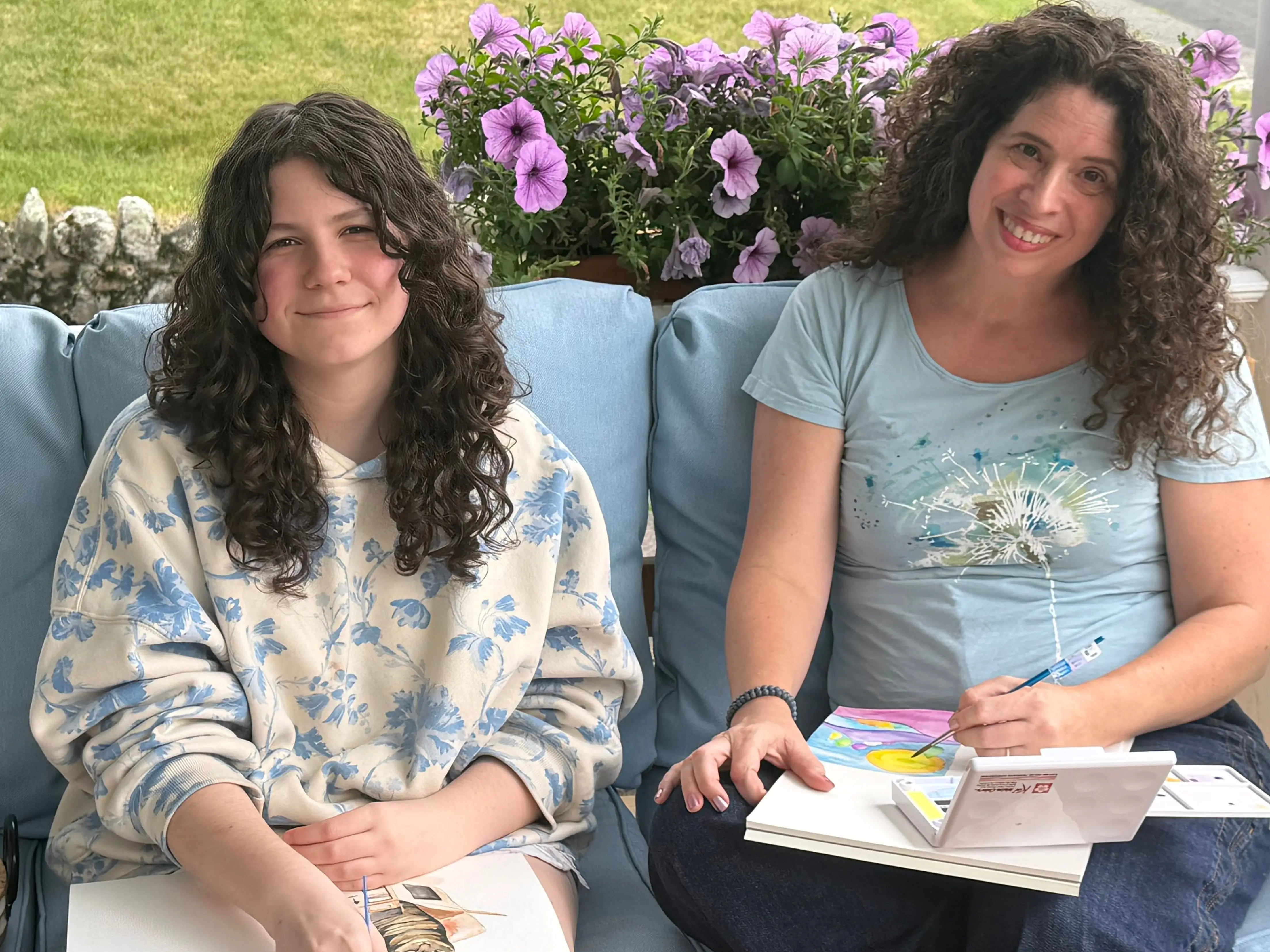 Two people sit on a blue outdoor couch with sketchbooks and art supplies beside purple flowers.