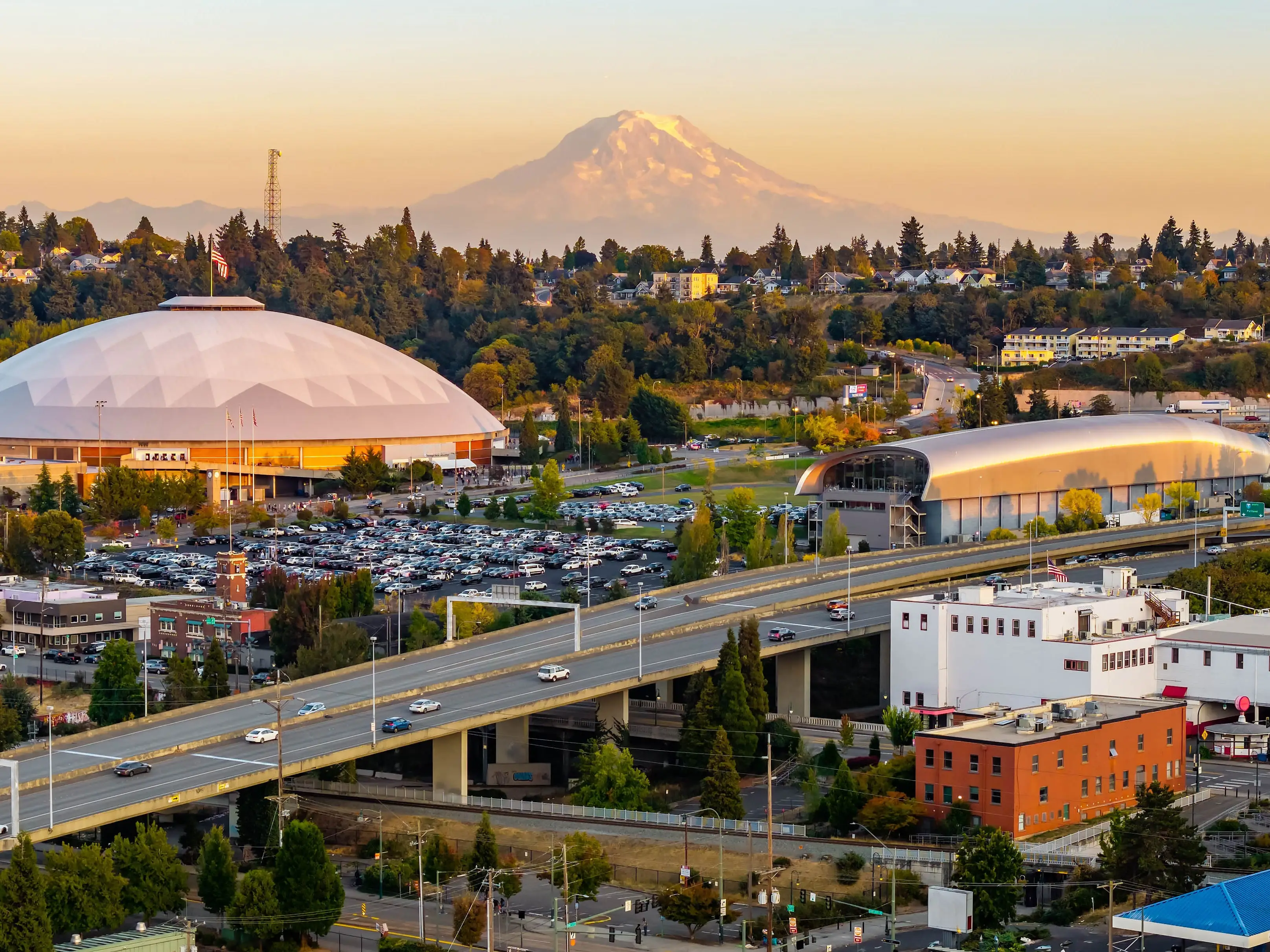 The Tacoma cityscape and its skyscrapers are dramatically framed against the backdrop of the towering, snow-capped Mount Rainier at sunset.