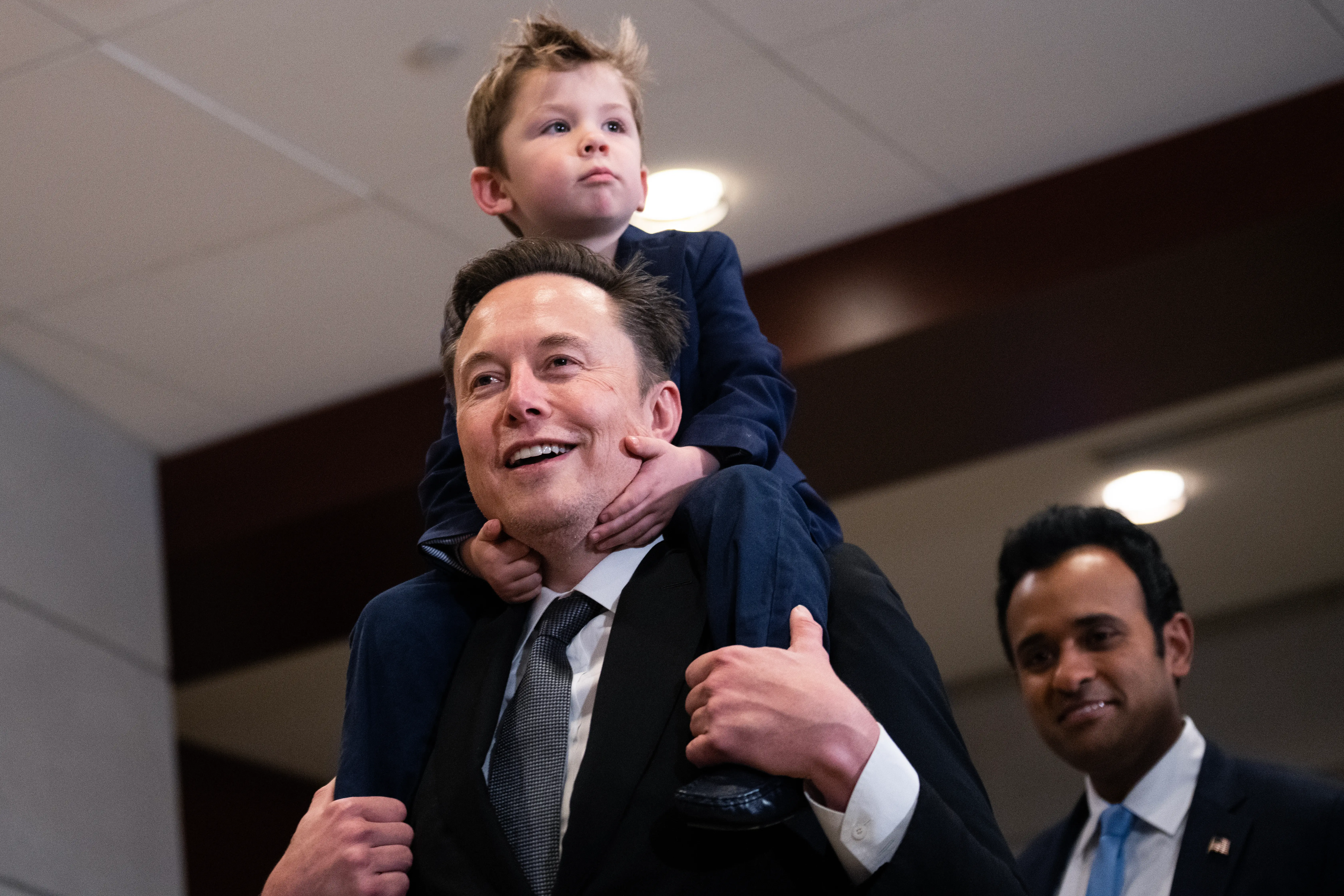 Elon Musk carries a child on his shoulders as Vivek Ramaswamy stands nearby in an indoor setting.