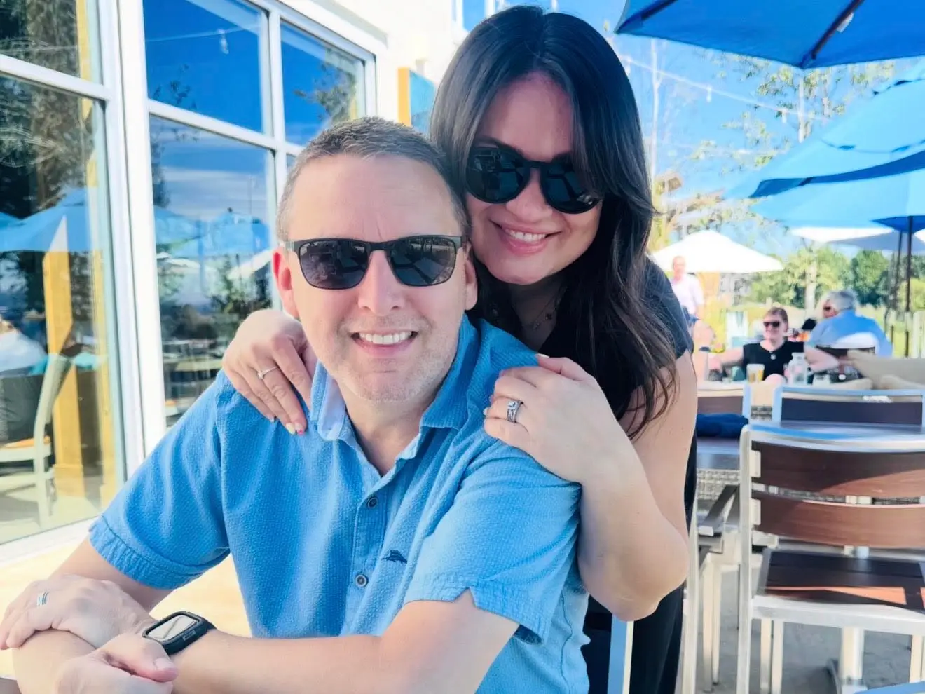 A woman hugs her husband from behind as he sits in a chair.