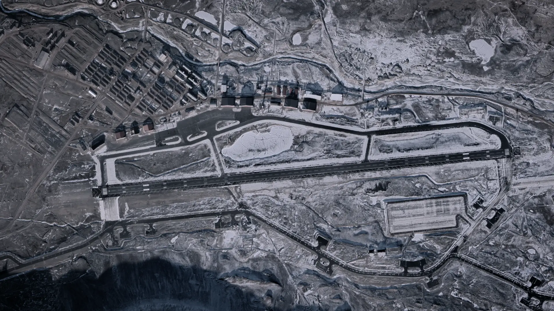A satellite image of a large runway on a rocky landscape with no vegetation and surrounded by a few dozen buildings.