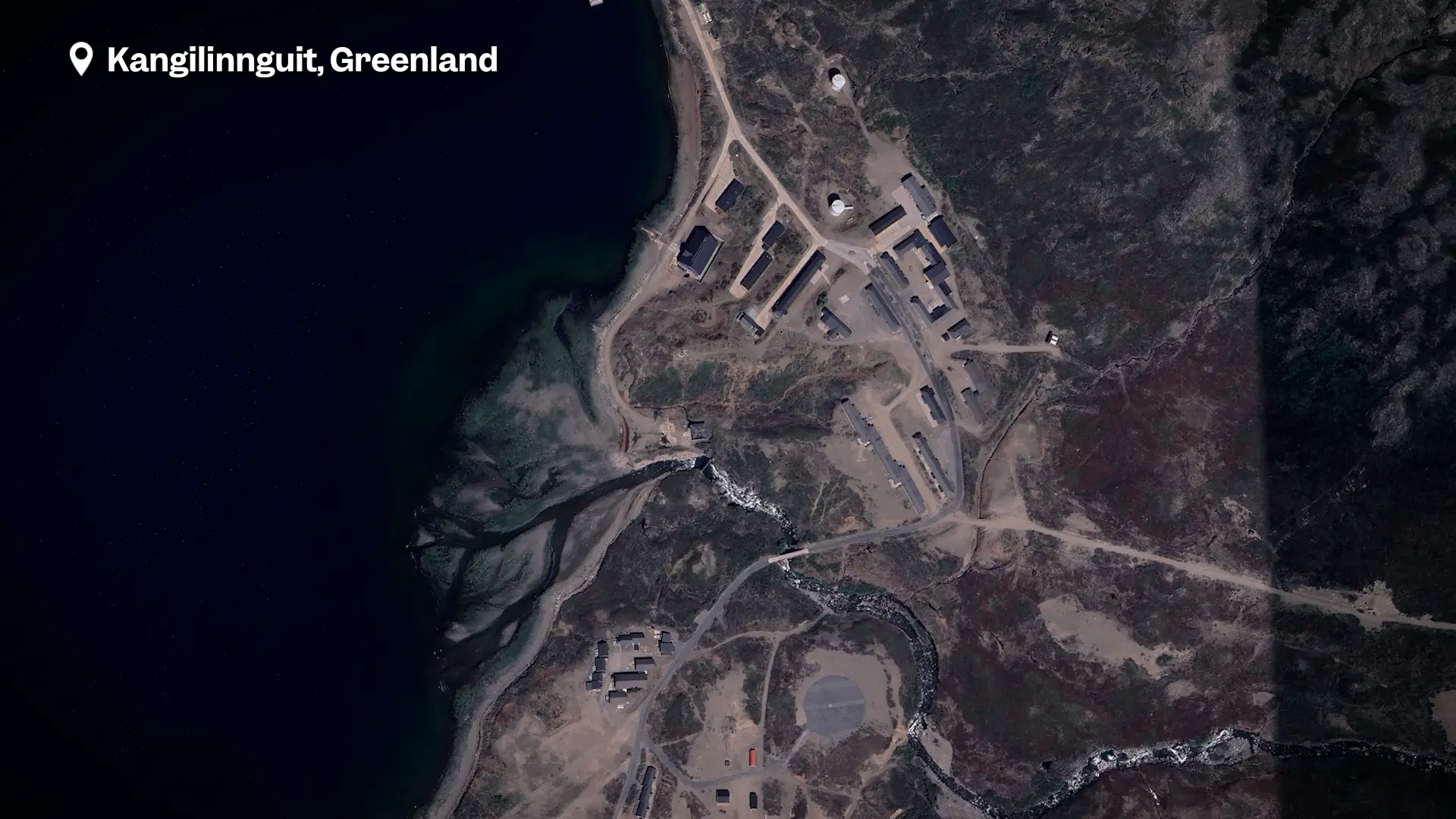 A satellite image of a coastal port with a couple dozen buildings among thin vegetation.
