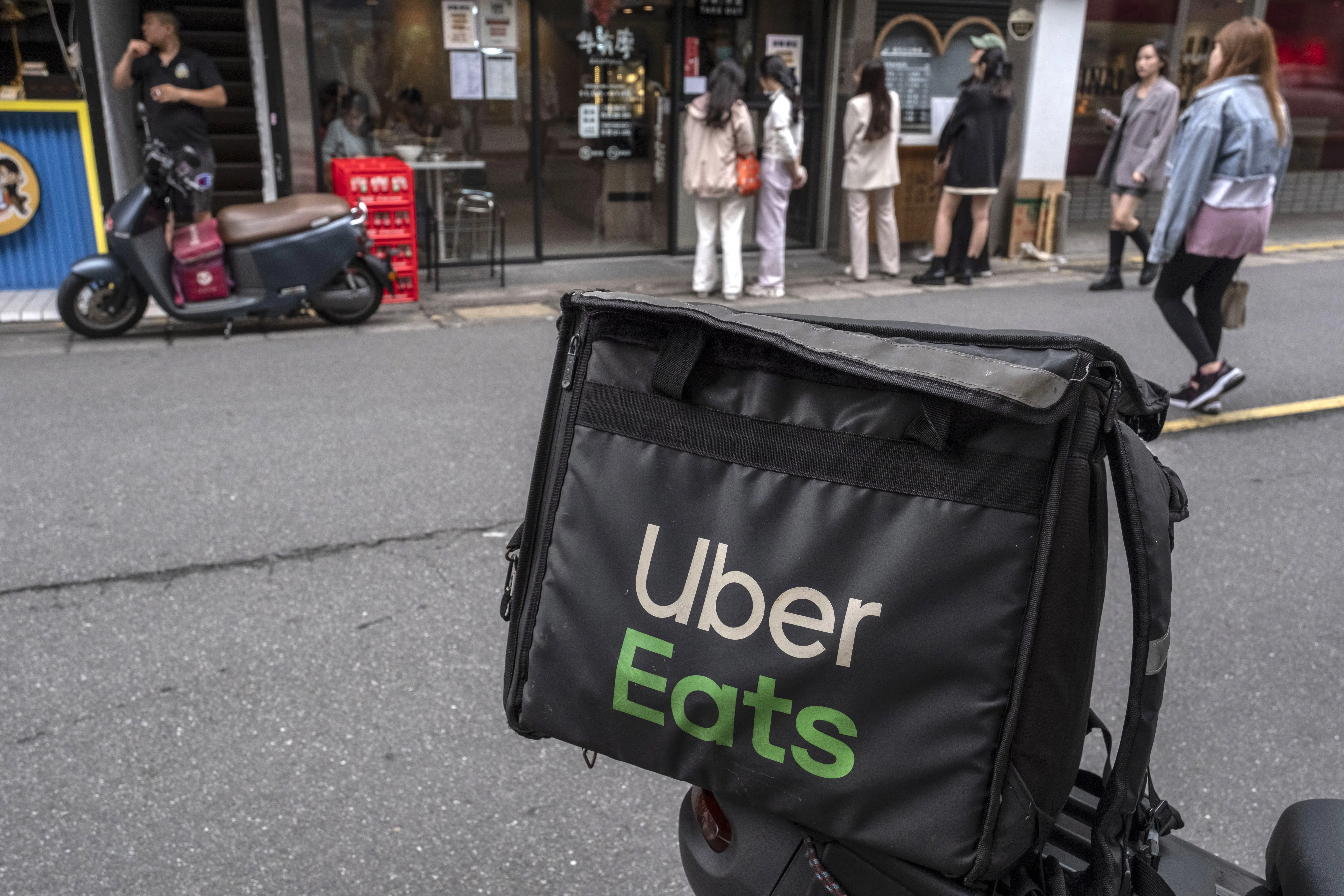 A black insulated bag sits on a moped as pedestrians cross a road in front of a storefront.
