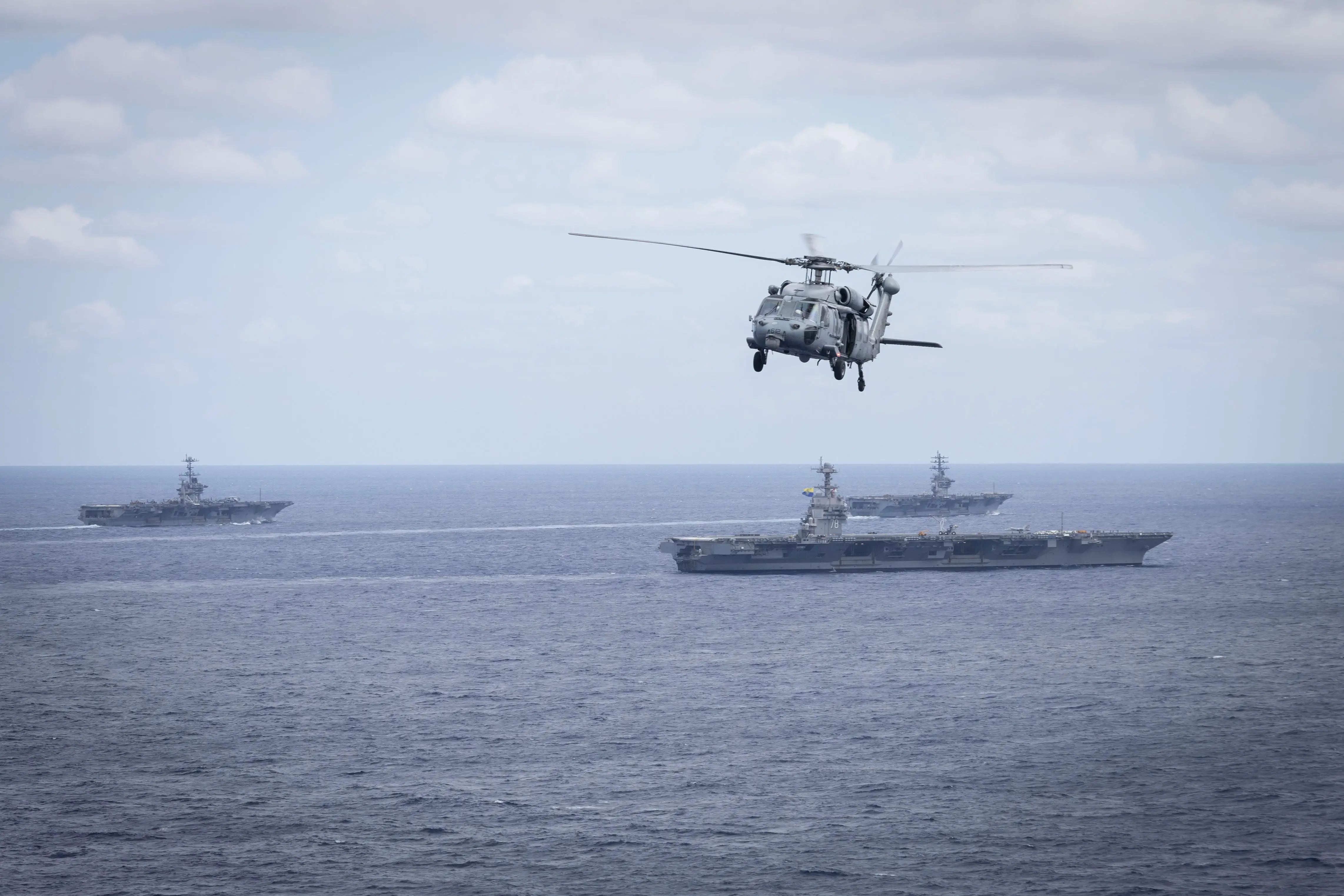 Three aircraft carriers sail in dark blue waters as a helicopter flies above in a cloudy blue sky.