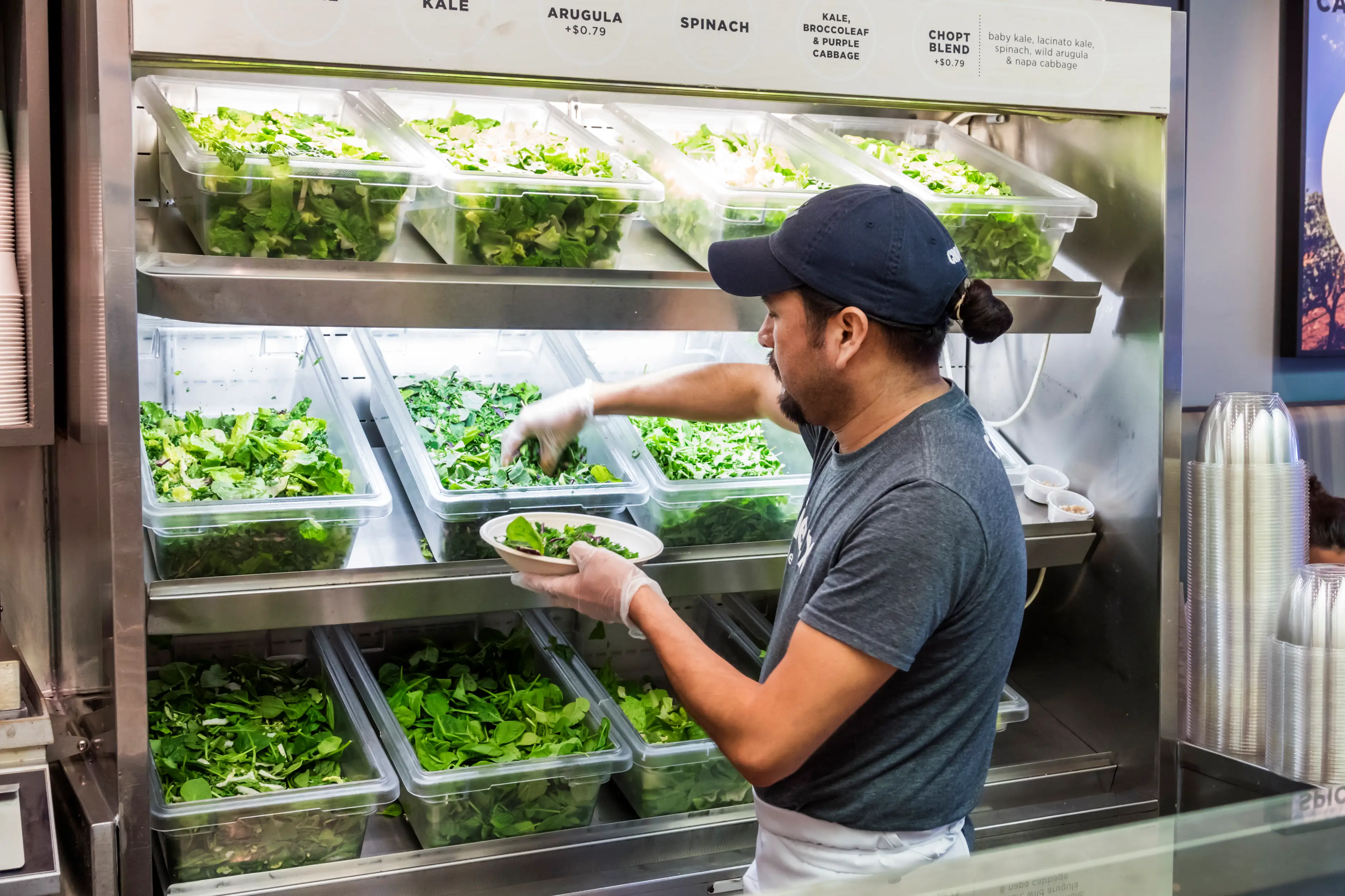 An employee at a Chopt restaurant picks leafy greens from a plastic container and places them into a bowl behind the counter while wearing a baseball hat.