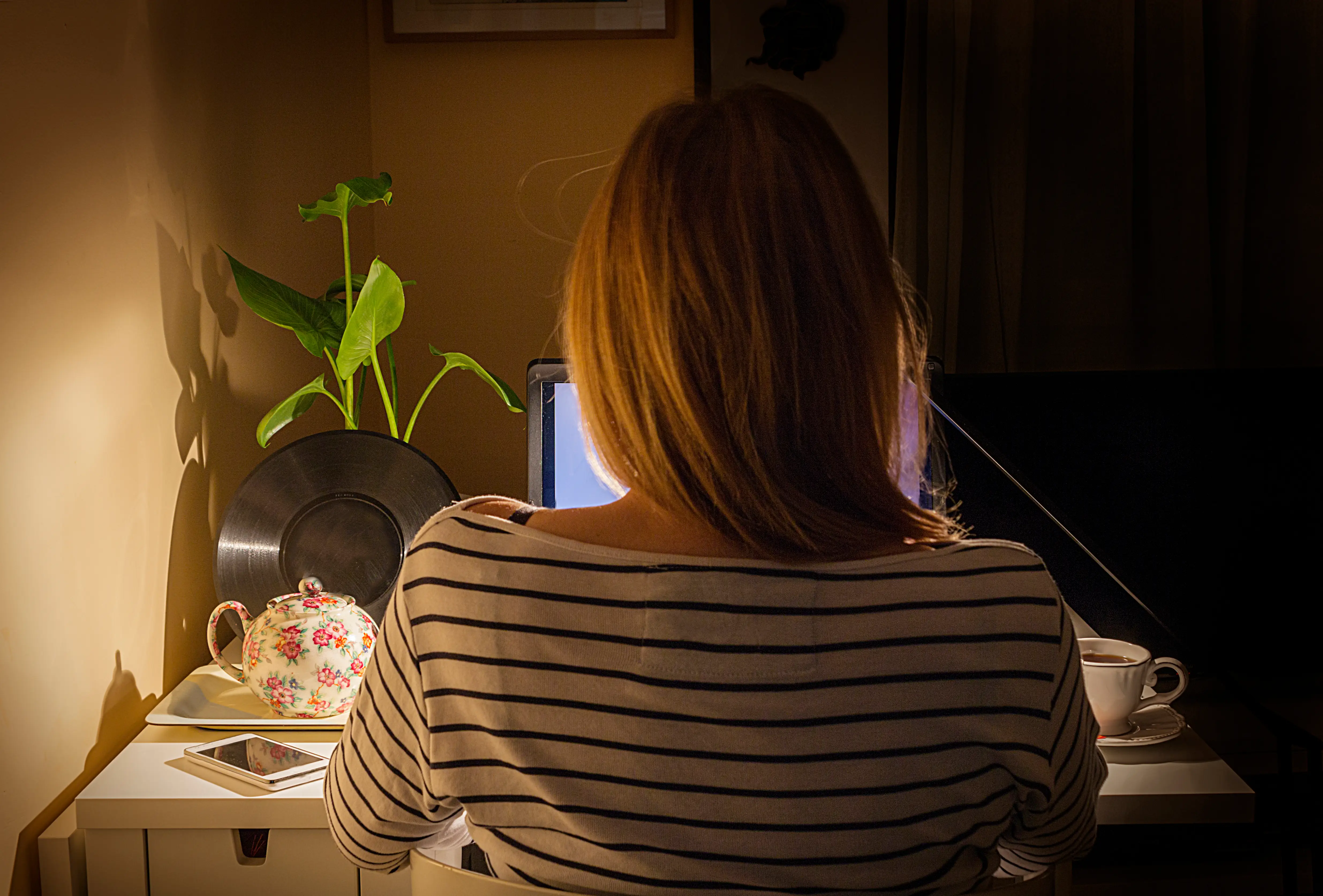 A woman from behind is working with a computer. It is night and the desk is illuminated by the light of a lamp