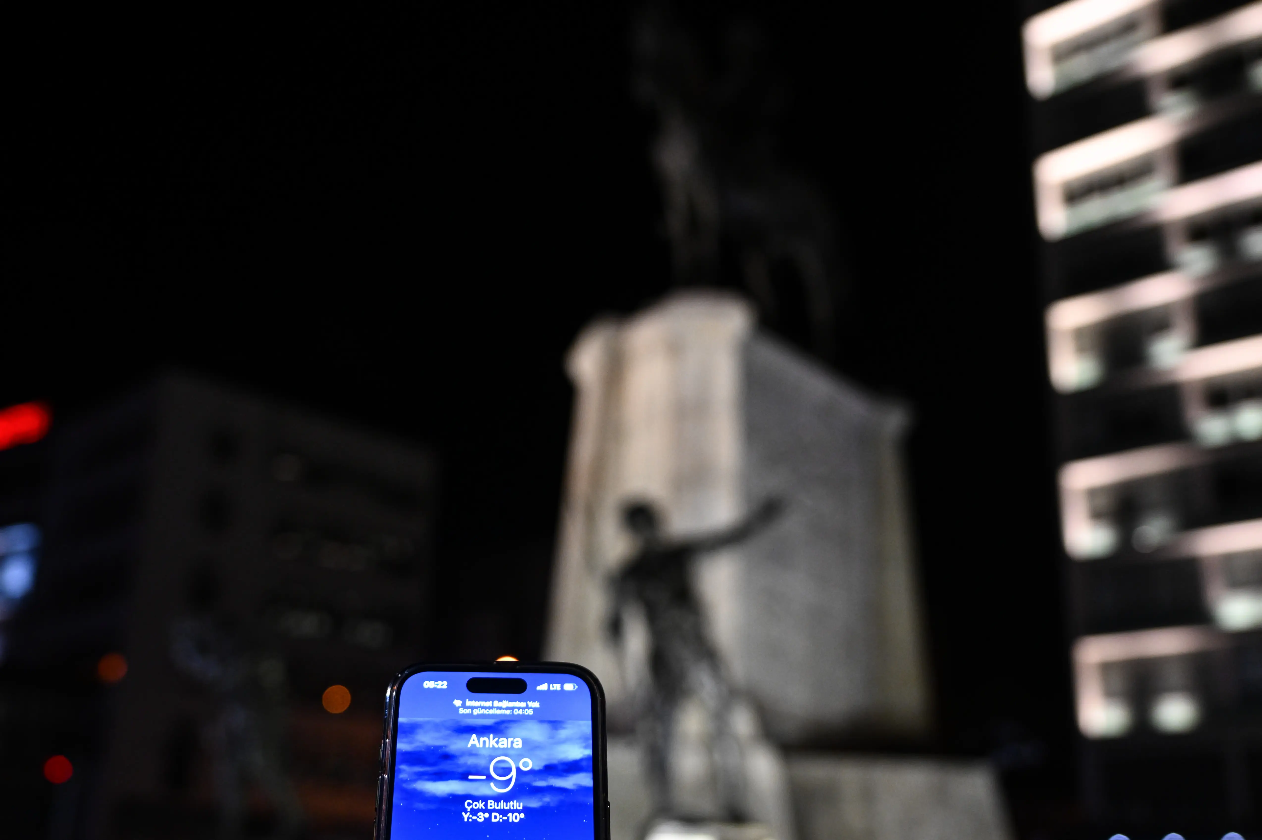 Smartphone weather screen shows Ankara at -9° at night, with a blurred illuminated monument and buildings behind it.