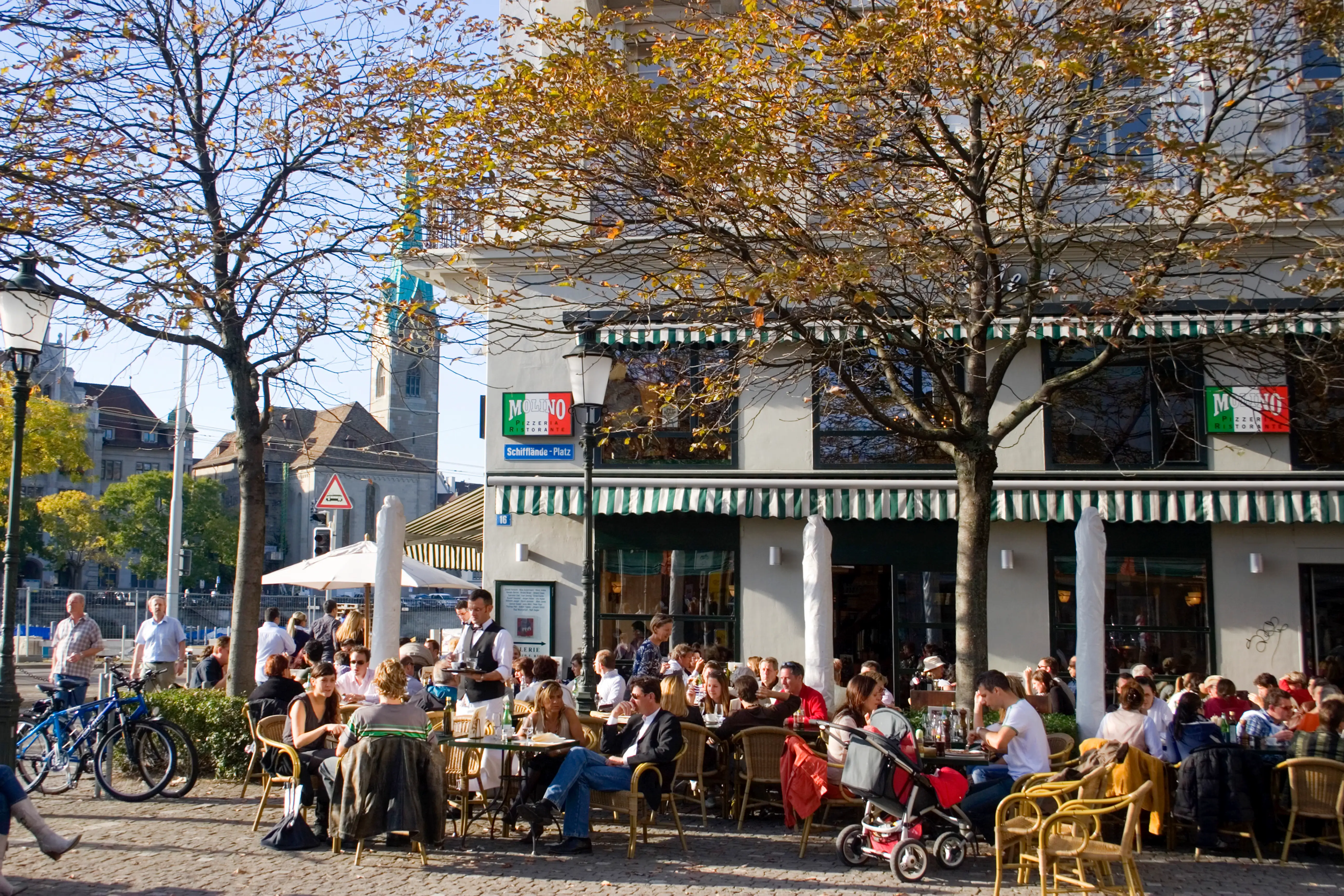 Crowded outdoor café terrace at Schifflände-Platz with diners, trees, bicycles, and striped awnings.
