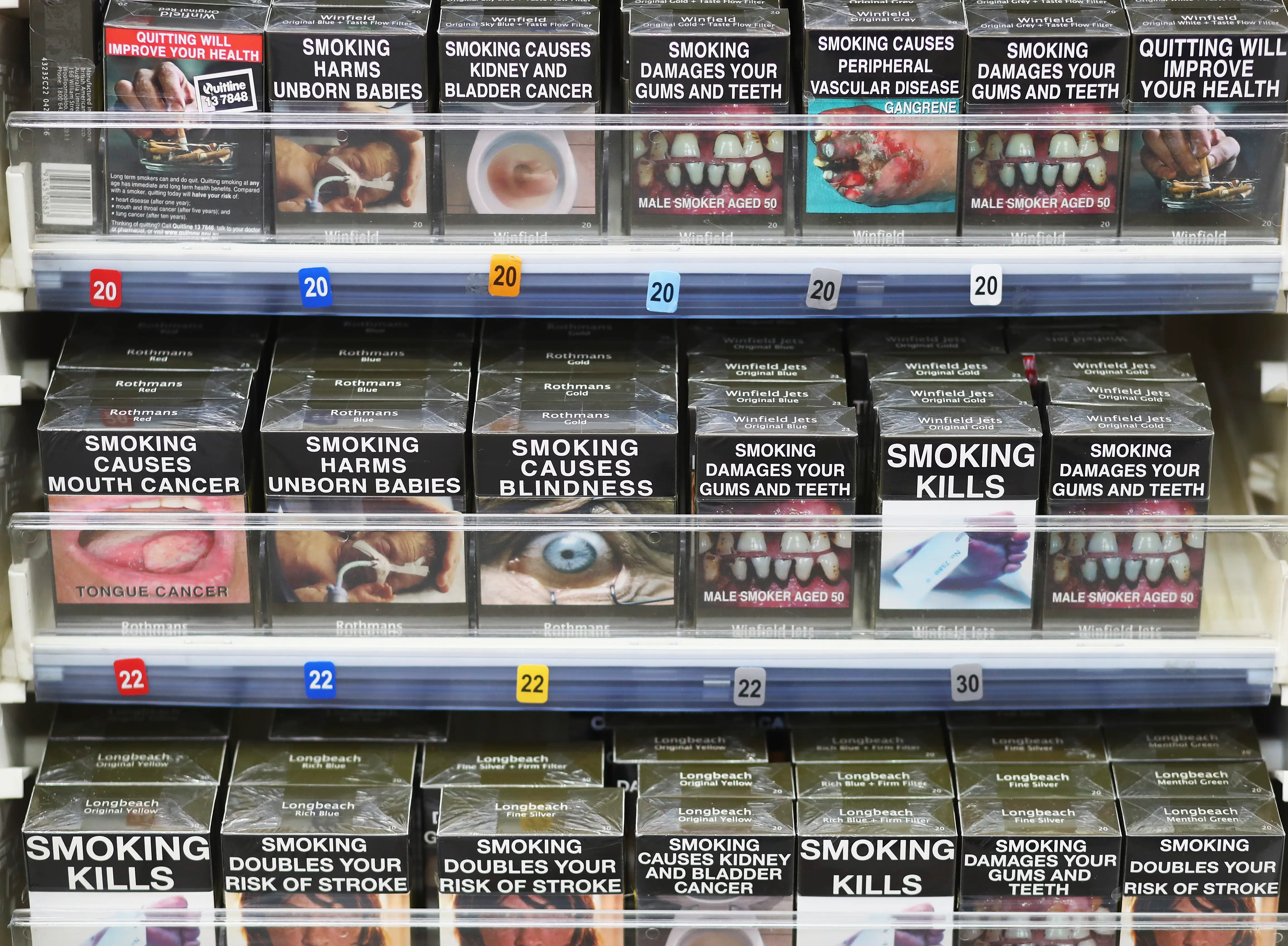 Cigarette packs with graphic health warnings are displayed on retail shelves with price labels.