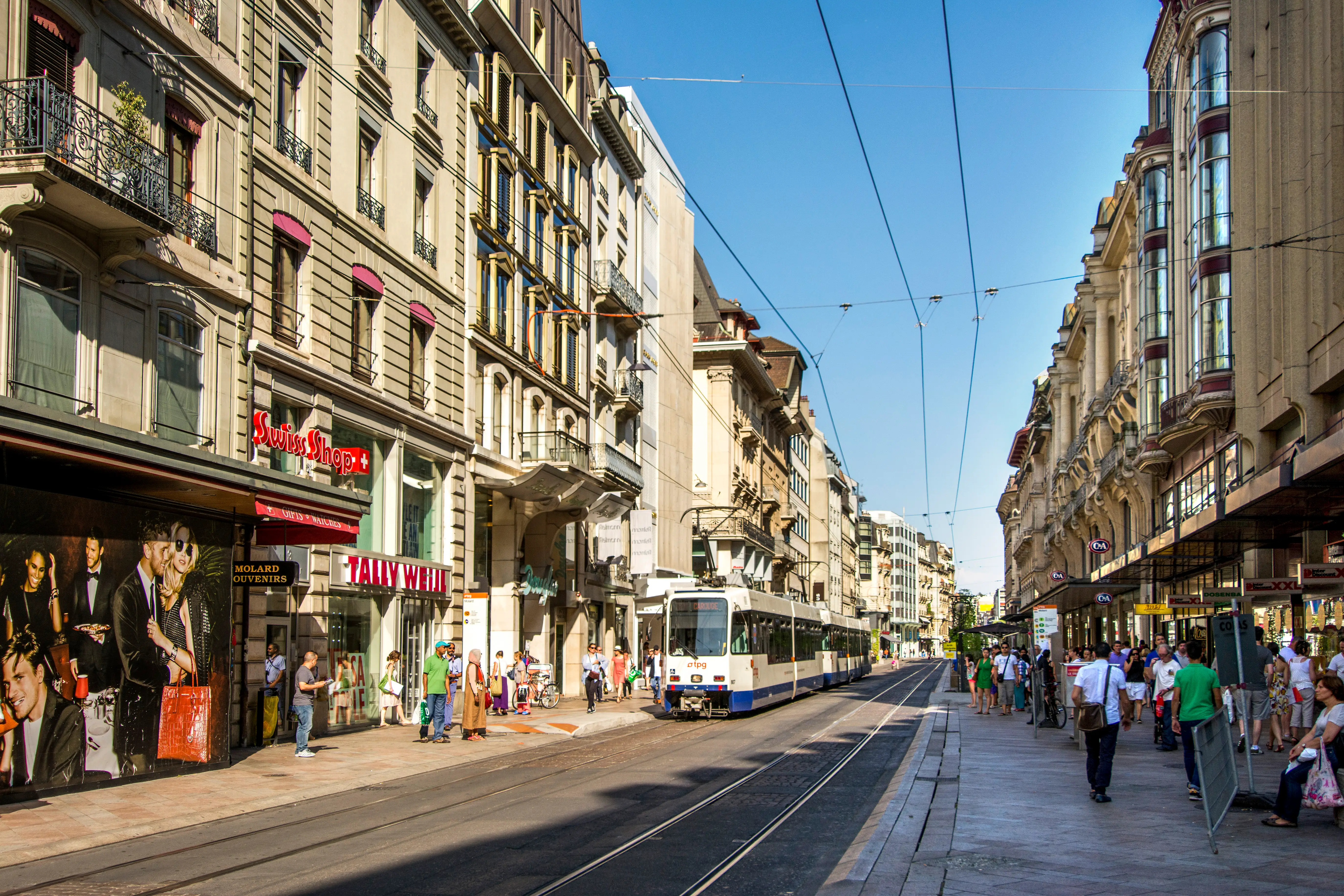 A tram travels along a busy shopping street lined with historic buildings, storefronts, pedestrians, and overhead wires.