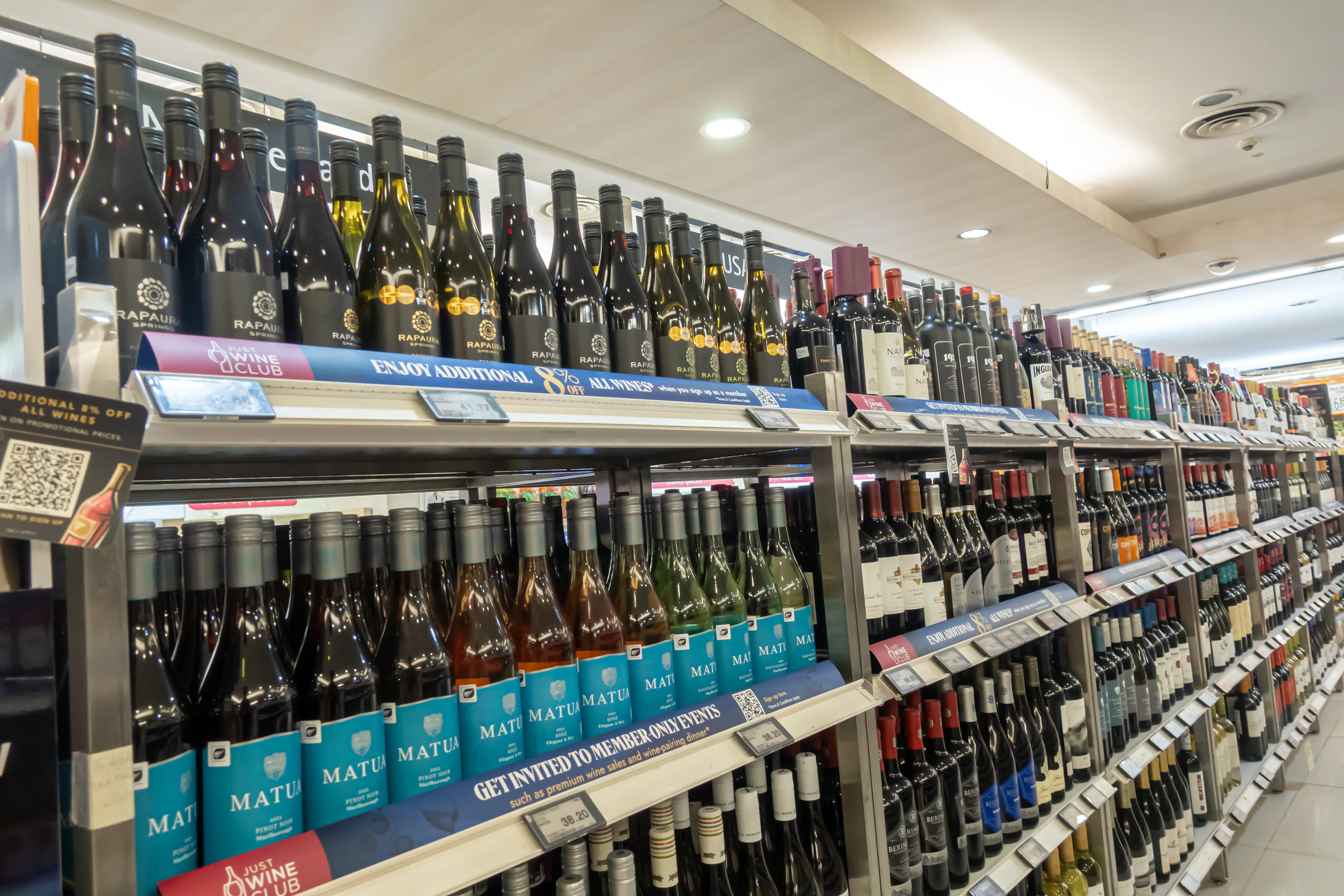 Rows of wine bottles line retail store shelves with promotional shelf signs for wine club discounts.