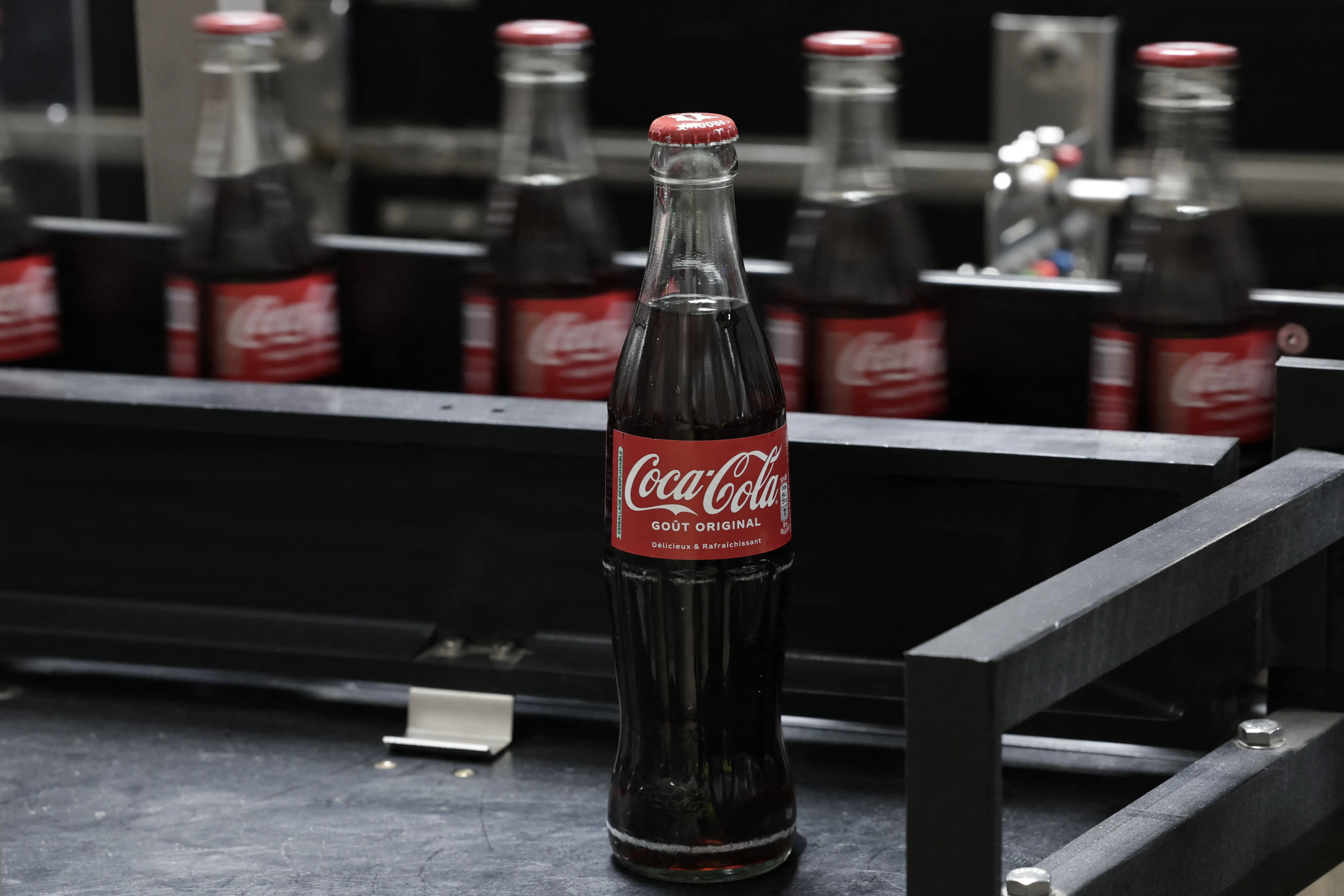 A Coca-Cola glass bottle stands on a bottling line with additional bottles blurred in the background.