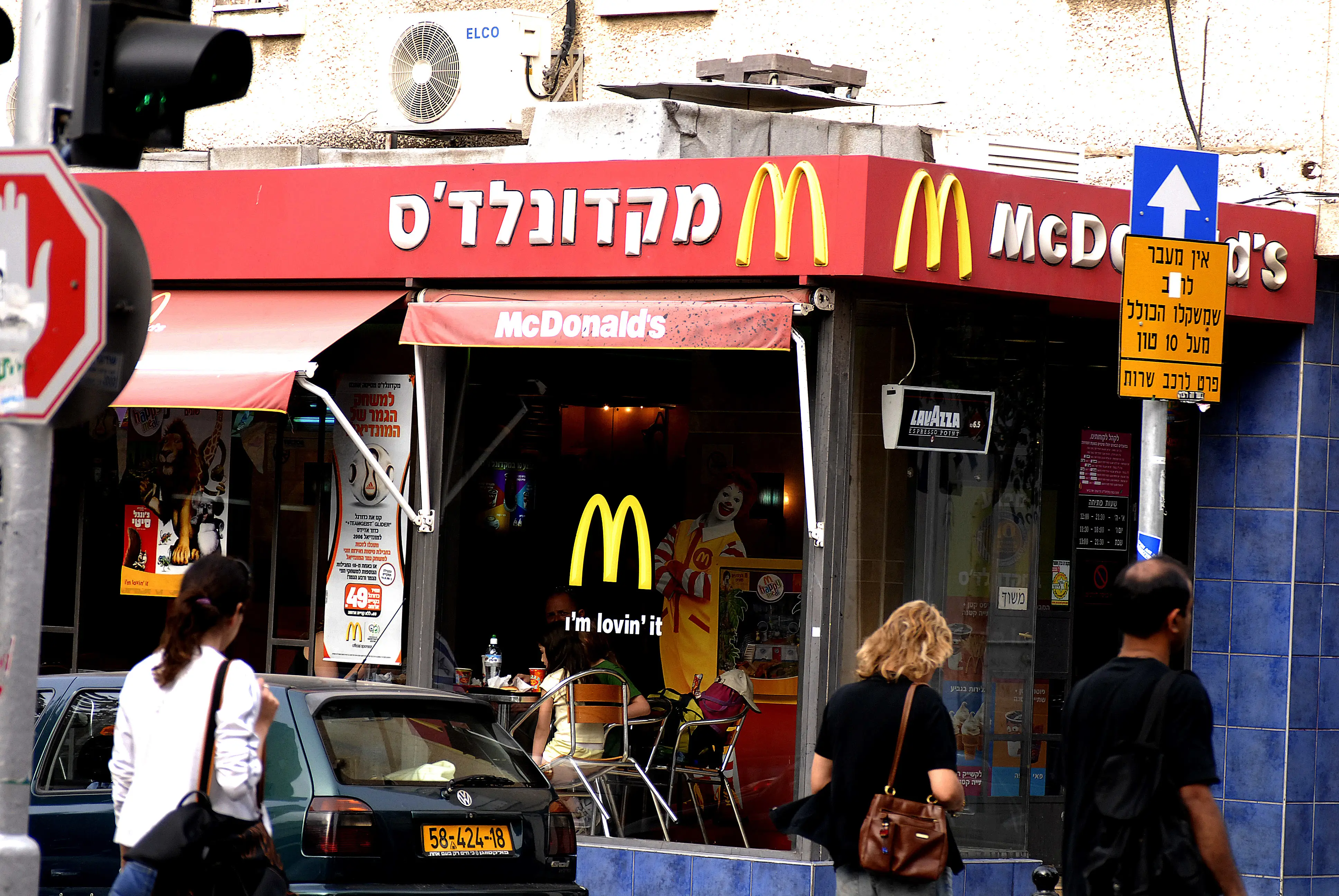 McDonald's storefront with Hebrew signage, golden arches logos, pedestrians, a parked car, and street signs outside.