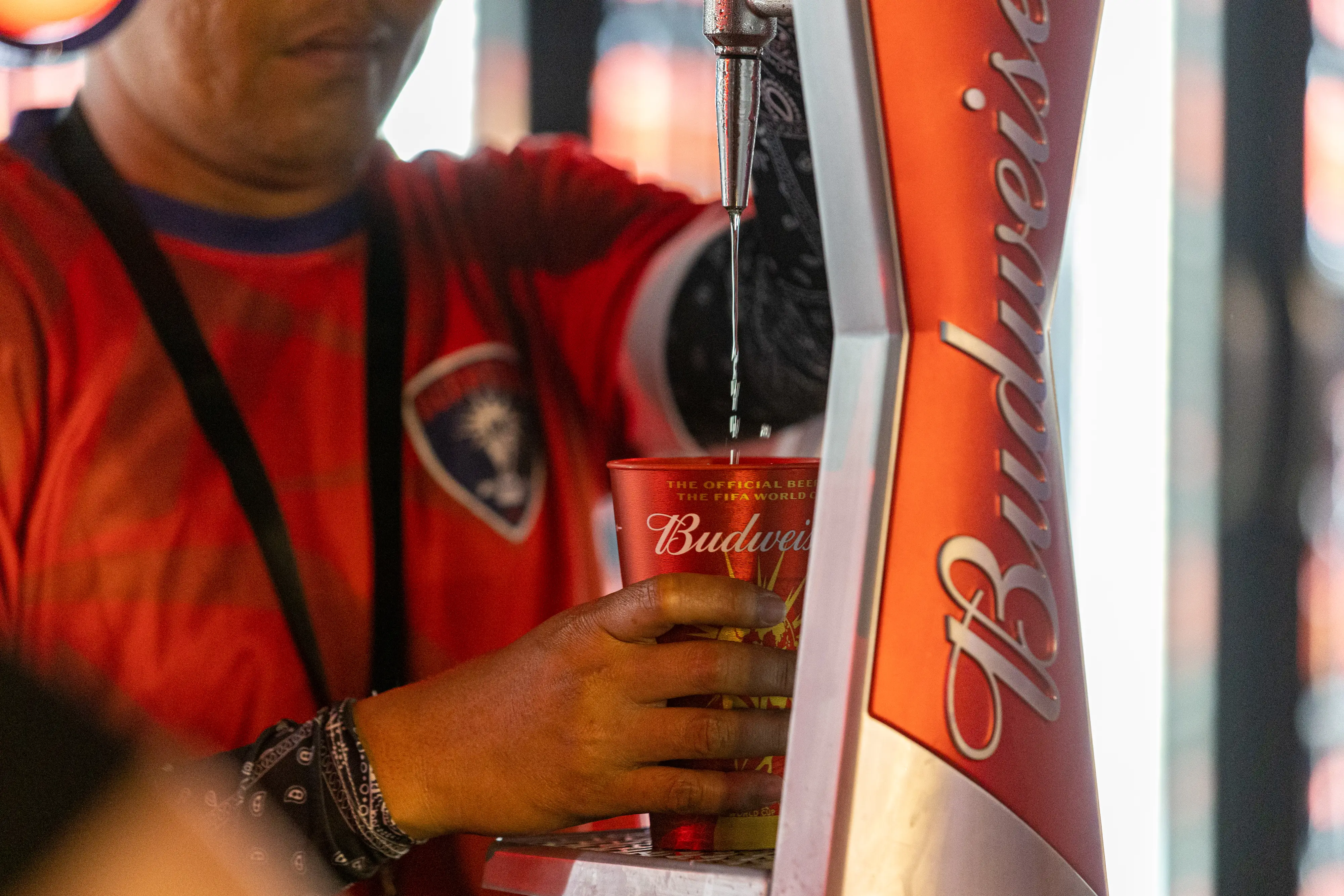 A person in a red jersey pours beer from a Budweiser tap into a branded plastic cup.