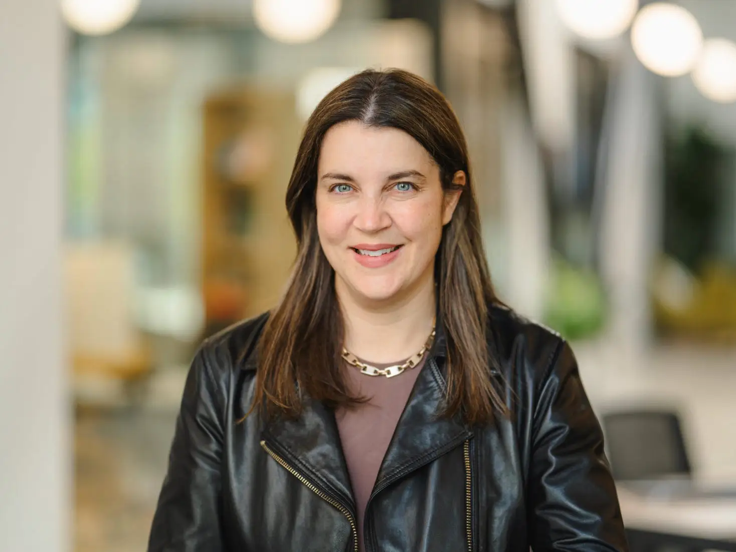 Woman in a black leather jacket poses for a portrait in a softly blurred office interior.