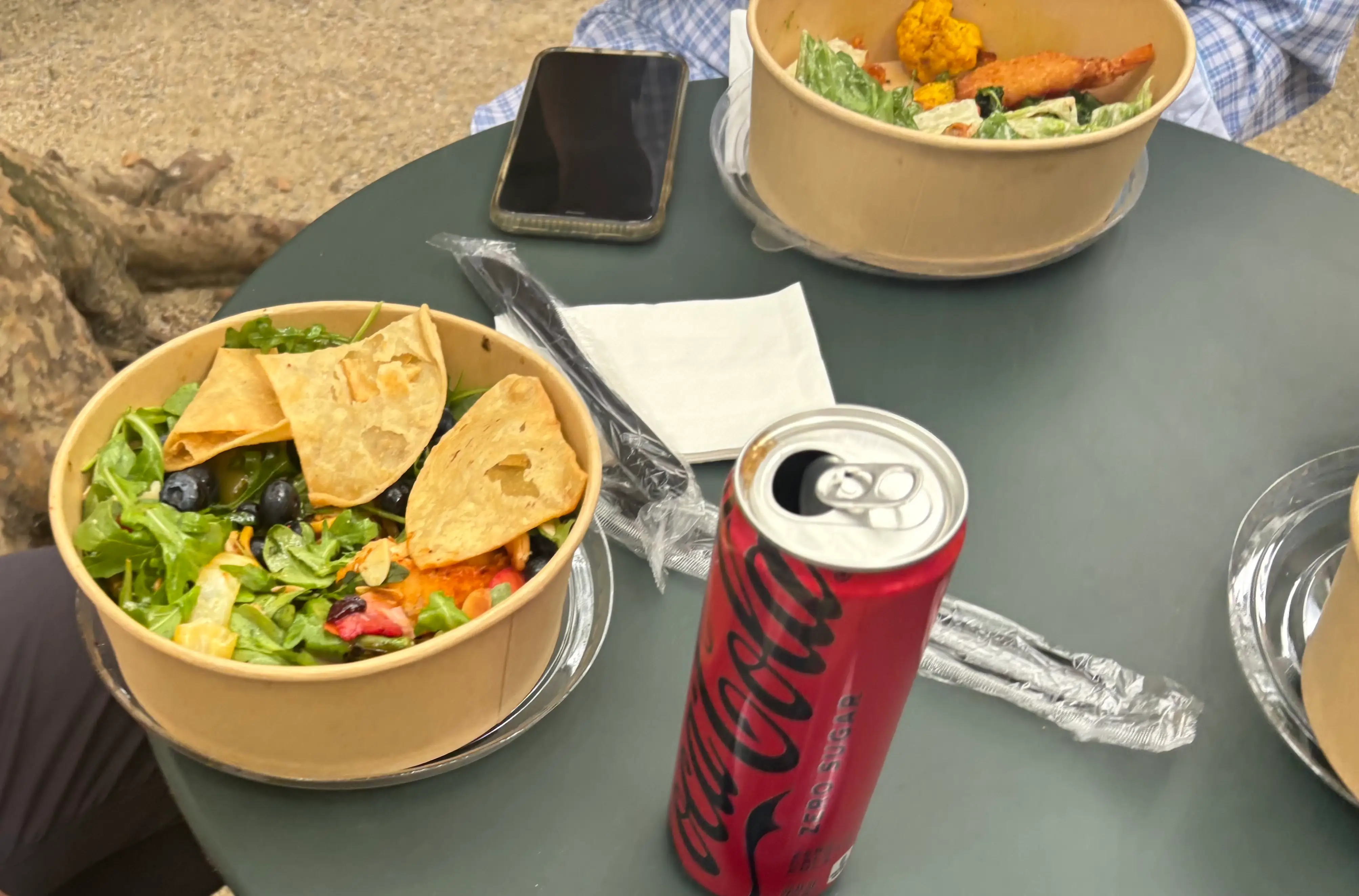 Two salad bowls, a Coca-Cola can, a smartphone, napkins and wrapped cutlery sit on an outdoor café table.