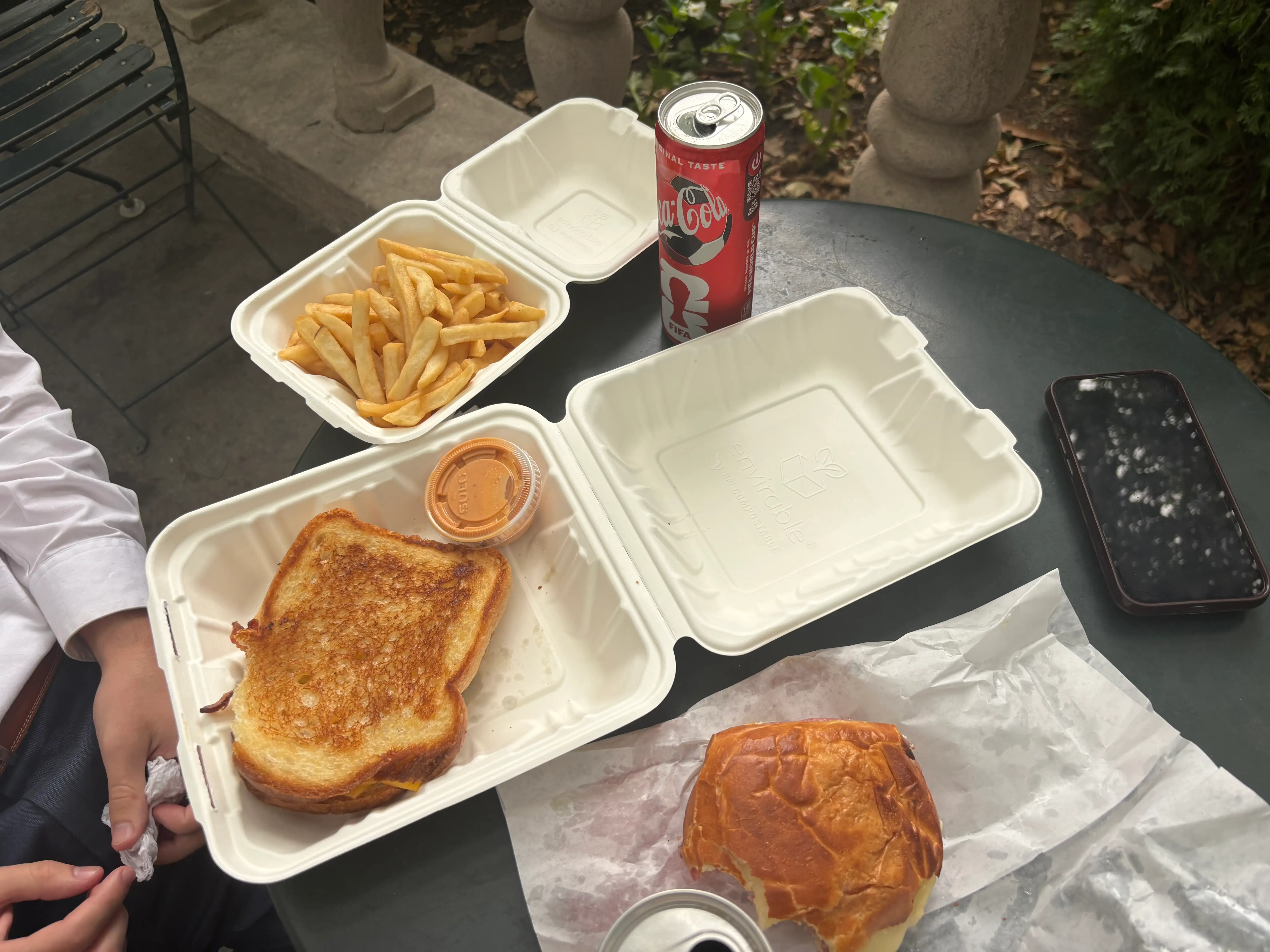 Takeout meals with fries, sandwiches, sauce, a Coca-Cola can, and a smartphone sit on an outdoor table.