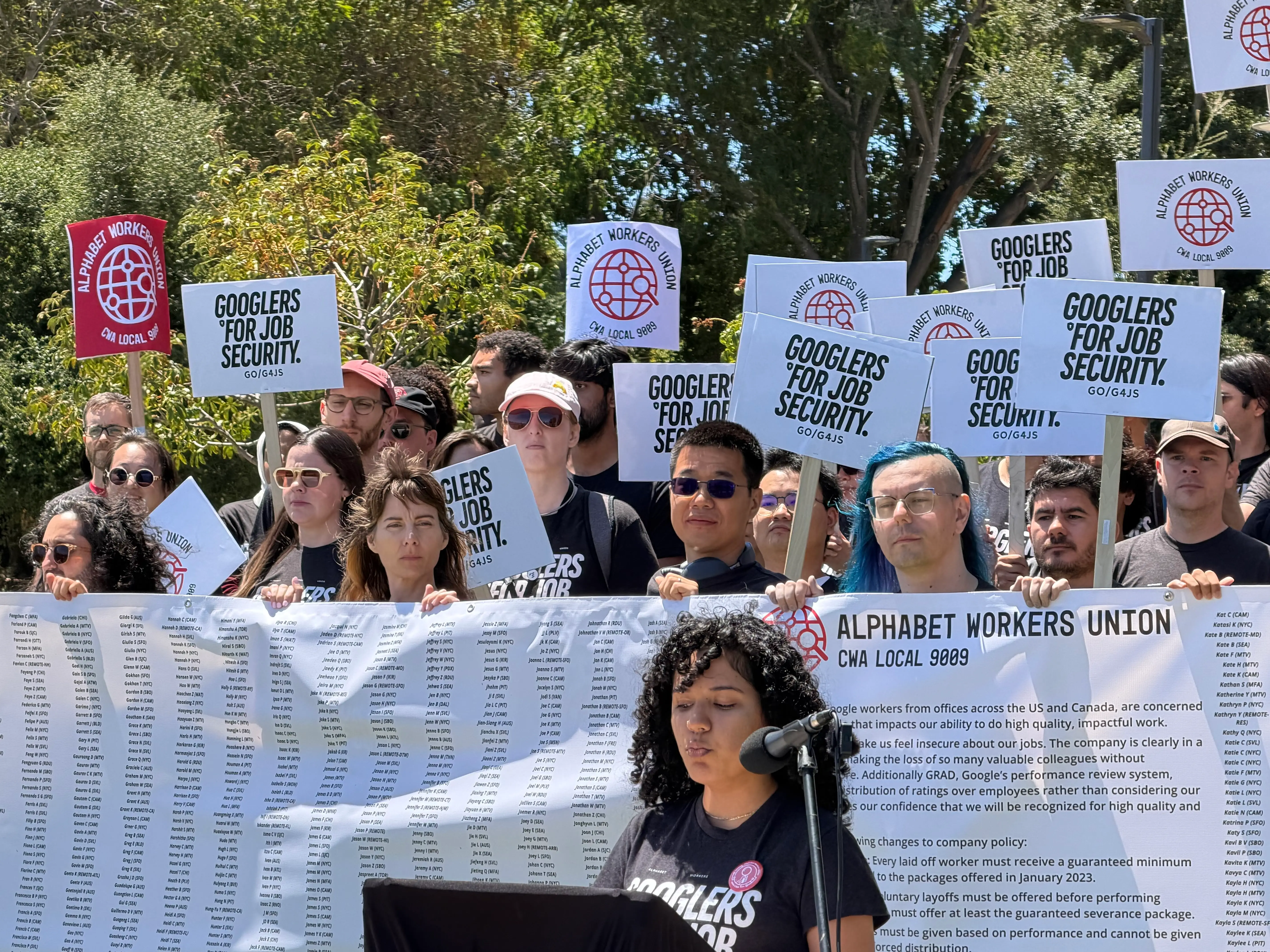 Crowd at an outdoor Alphabet Workers Union rally holds “Googlers for Job Security” signs behind a speaker.