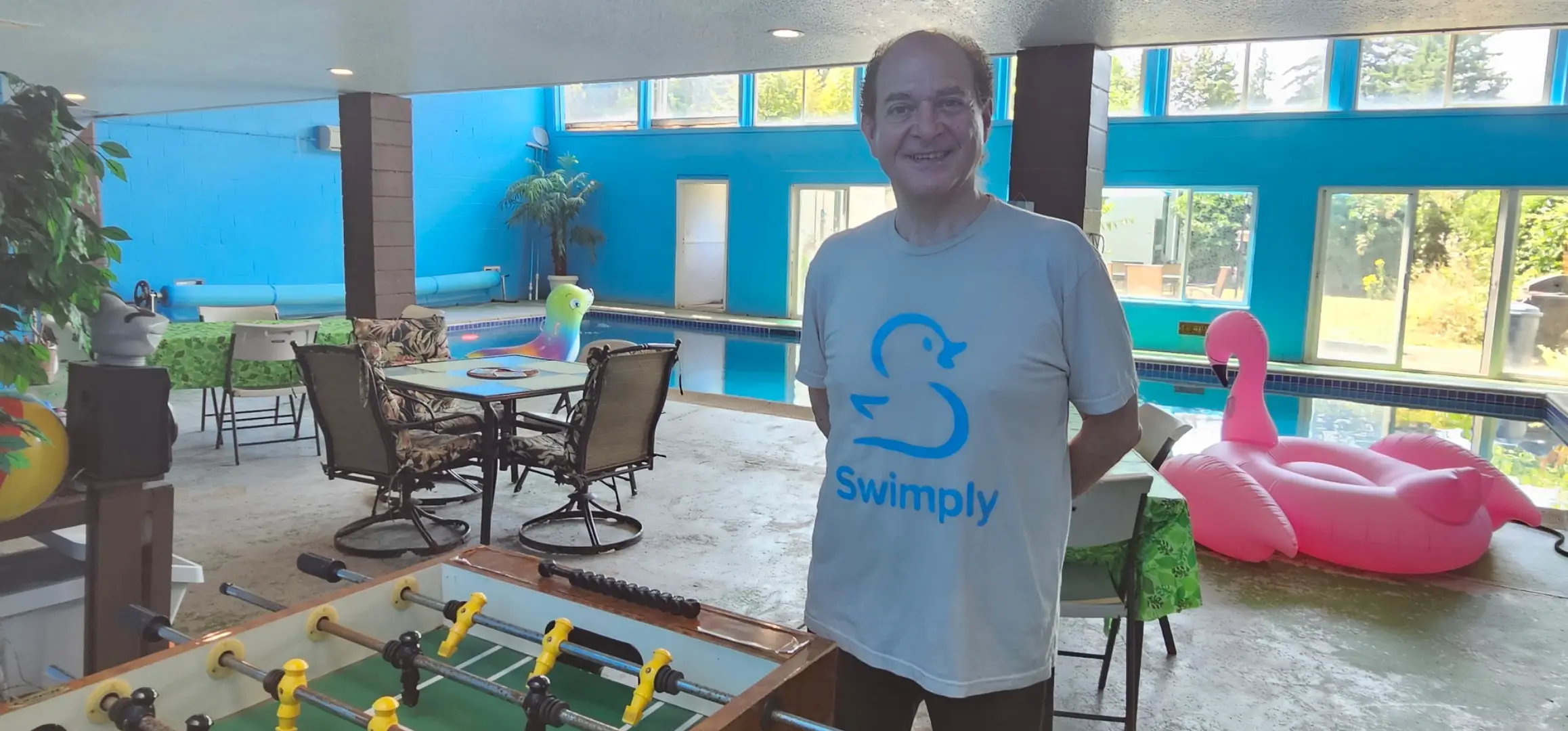Person wearing a Swimply T-shirt stands beside a foosball table in an indoor pool room.
