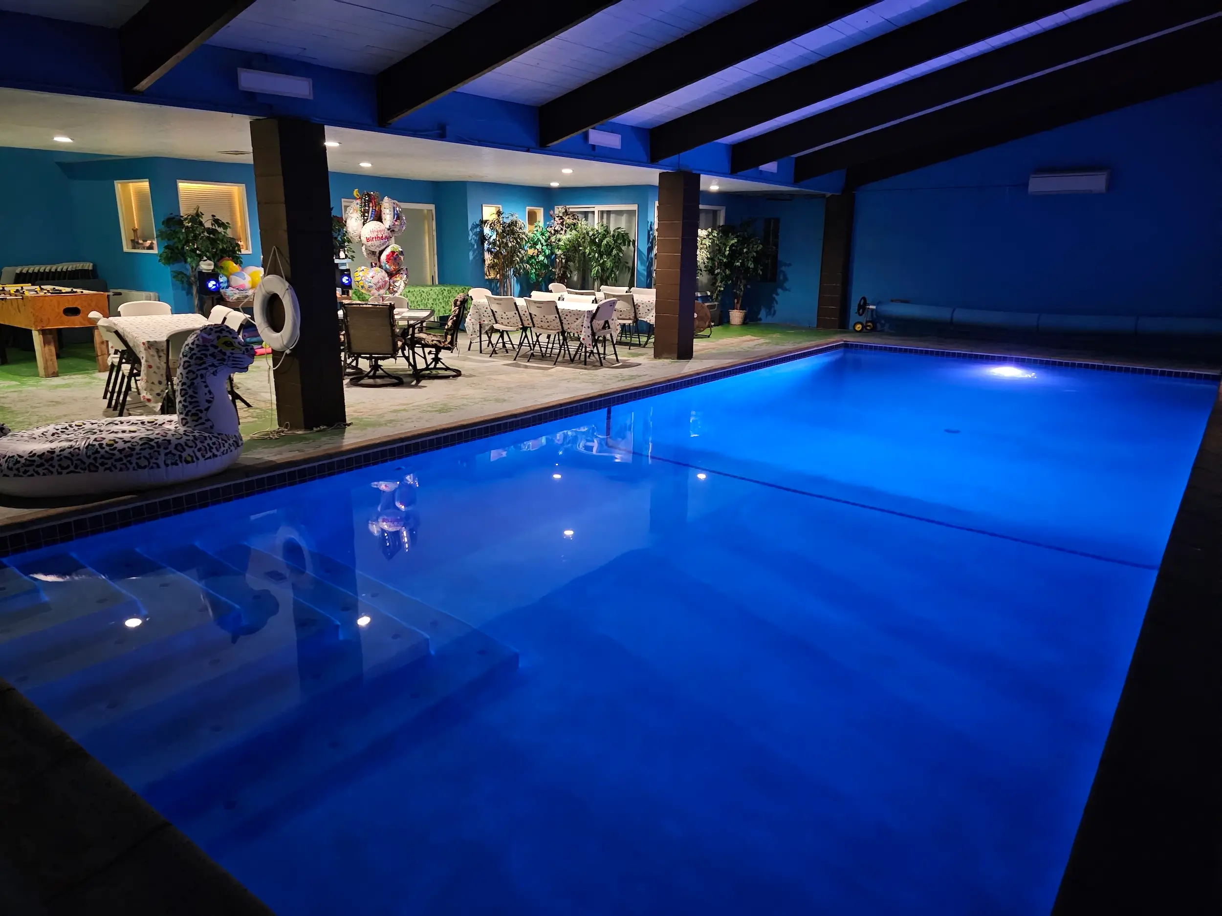 Covered swimming pool glows blue beside a decorated patio with tables, chairs, balloons, and party seating.