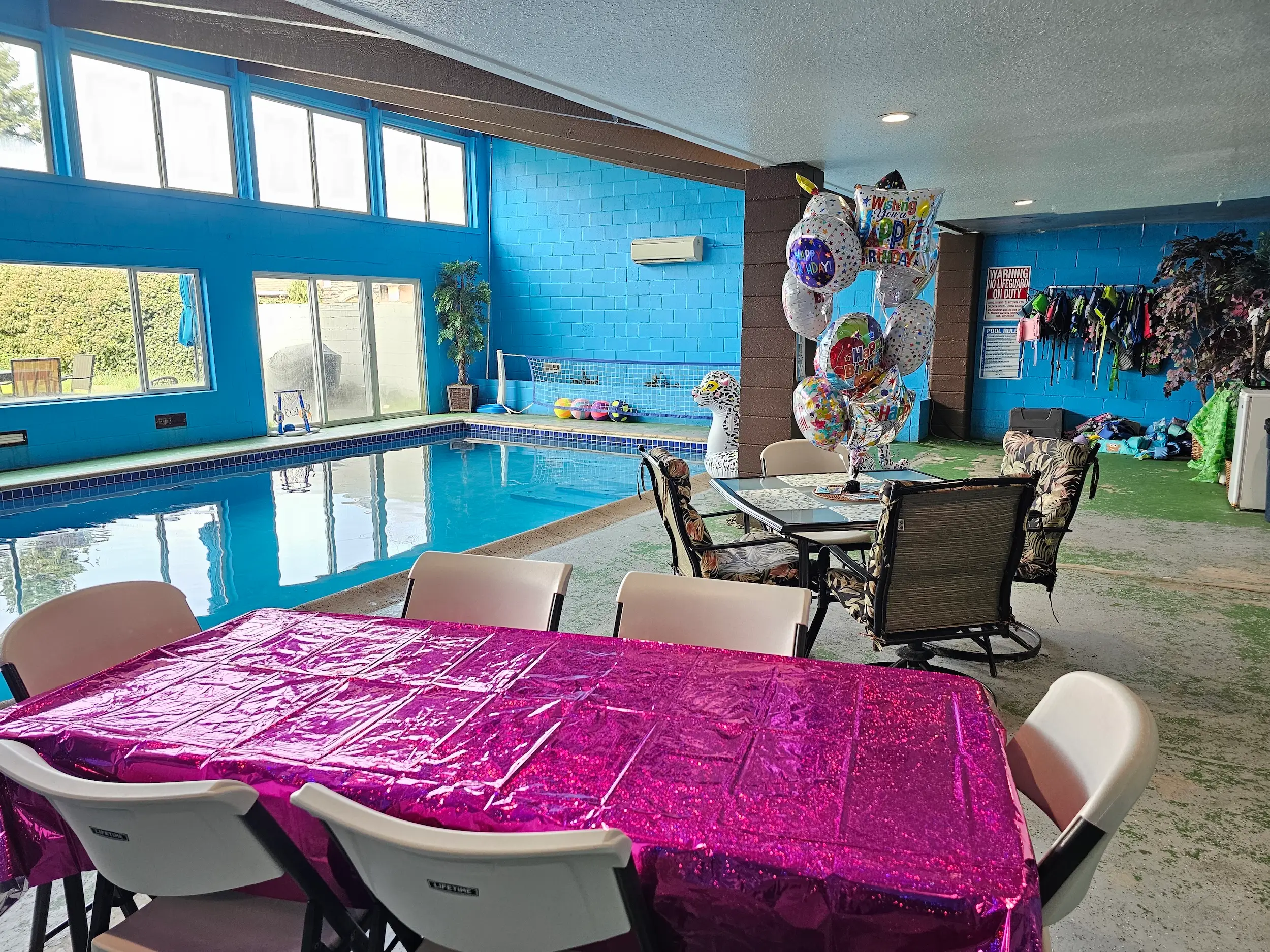 Indoor pool room decorated for a birthday party with balloons, folding chairs, and a pink tablecloth.