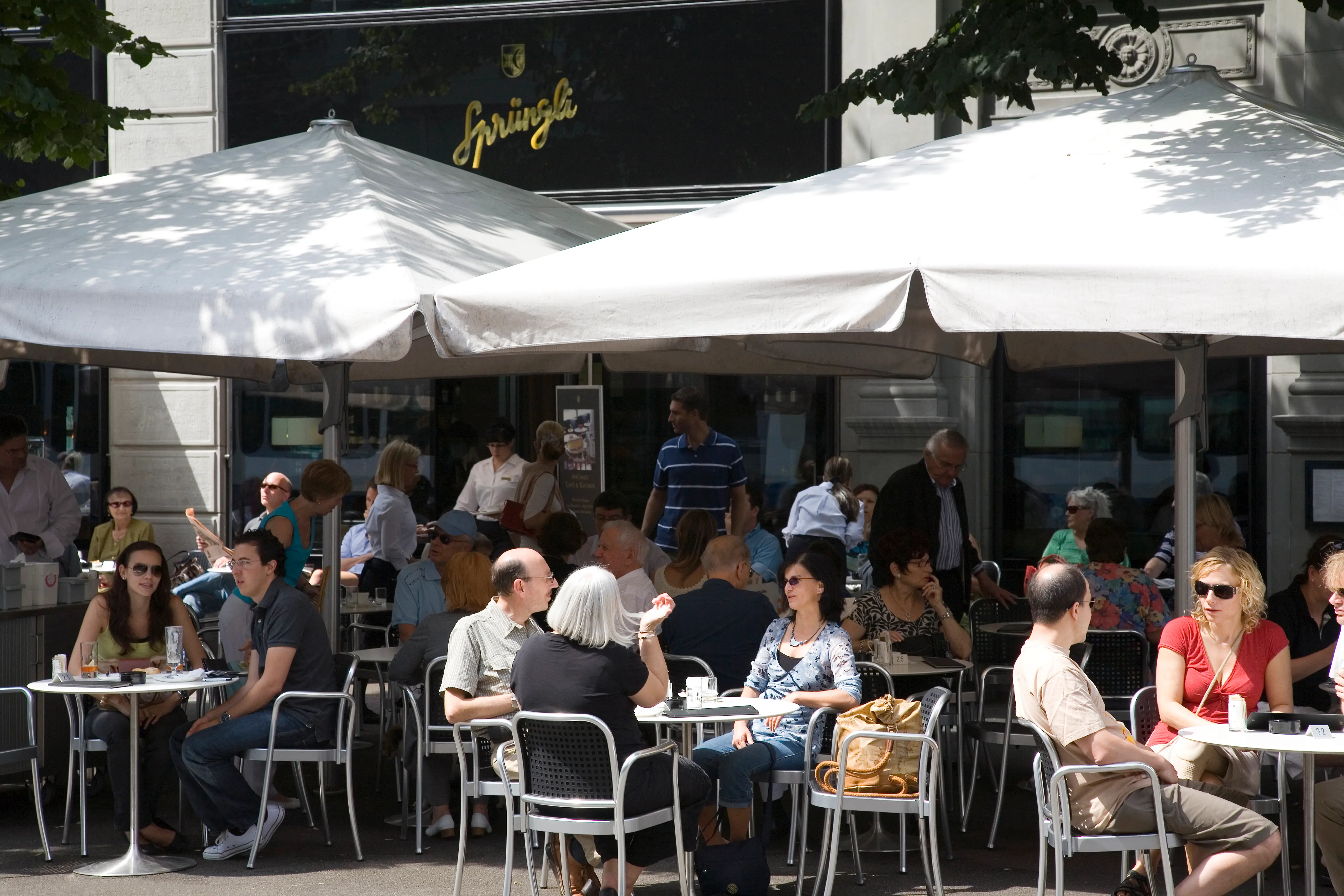 Patrons sit at outdoor café tables under large white umbrellas in front of a Sprüngli storefront.