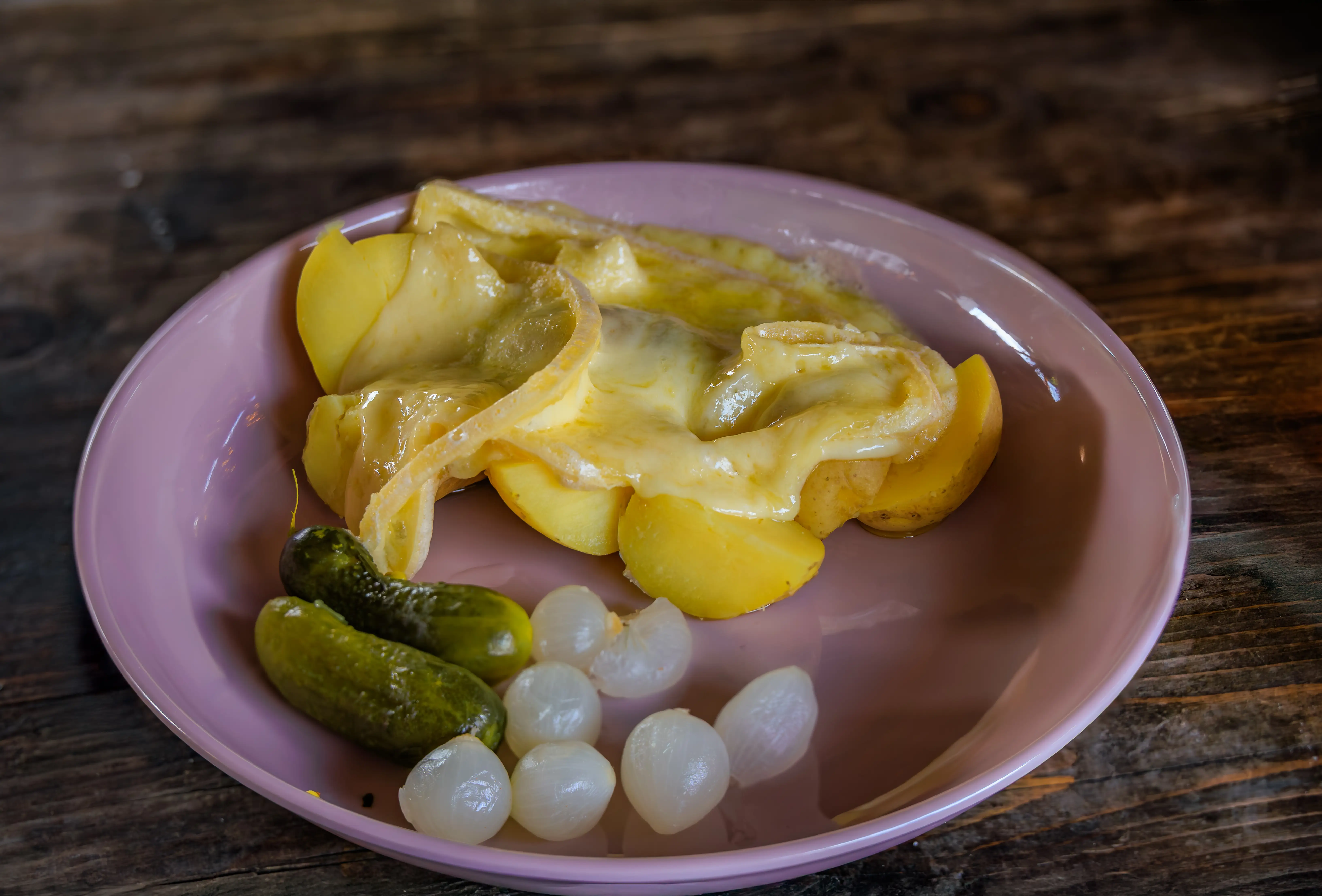 Melted cheese covers sliced potatoes on a pink plate with pickles and small onions on a wooden table.