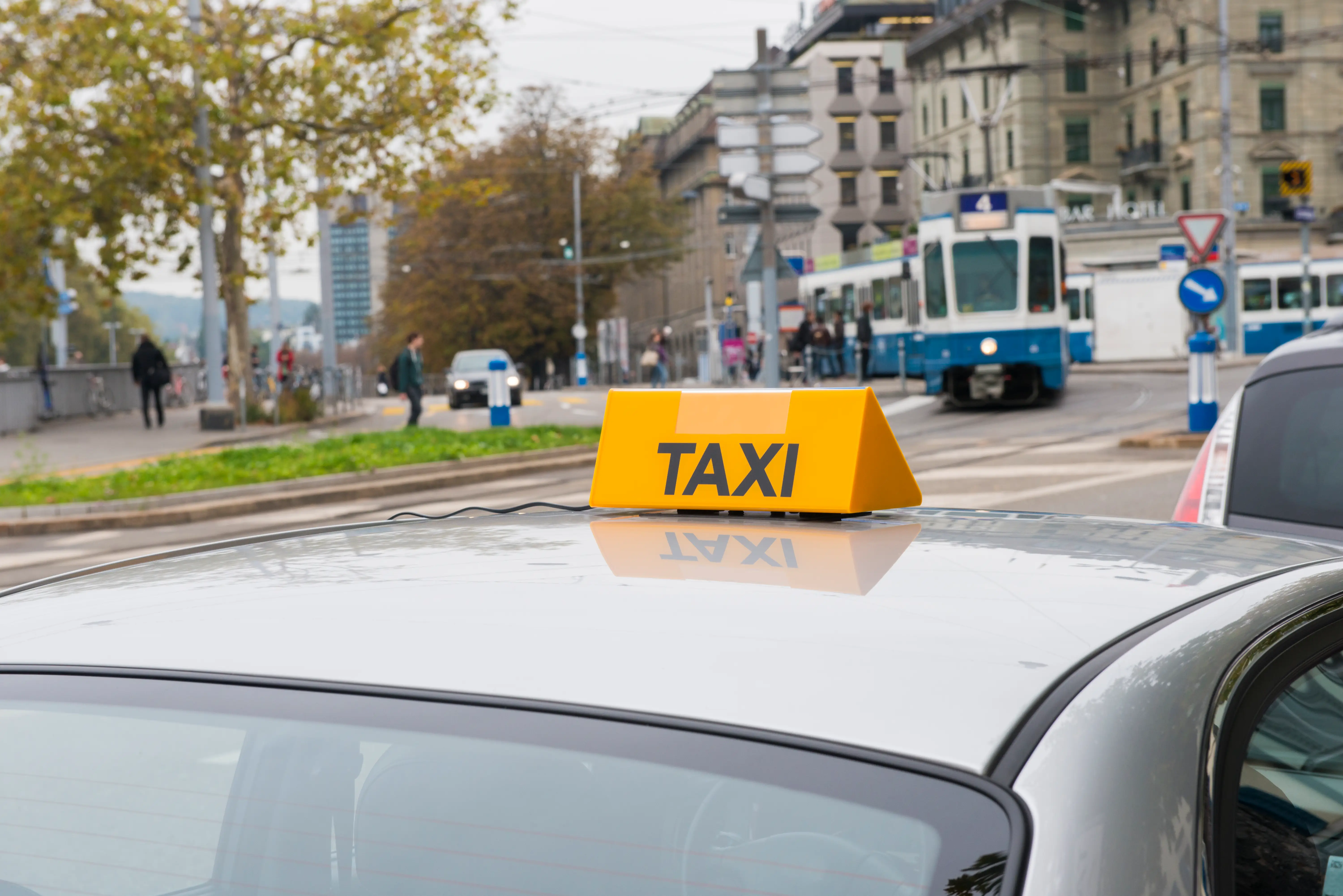 Yellow TAXI roof sign on a car with a blue-and-white tram and city street in the background.