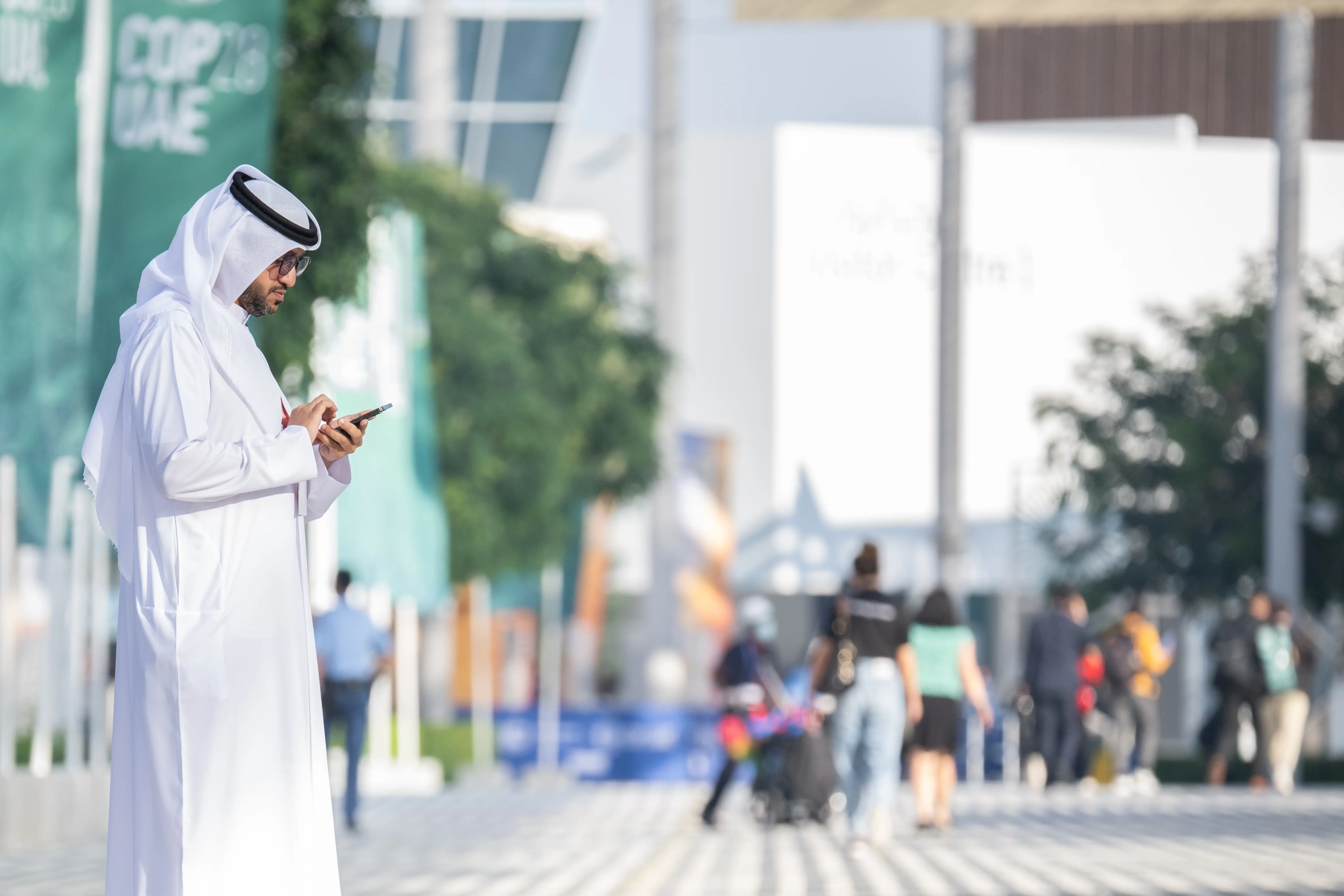 Person in white traditional Gulf clothing uses a smartphone beside a walkway with blurred Expo 2020 UAE signage.