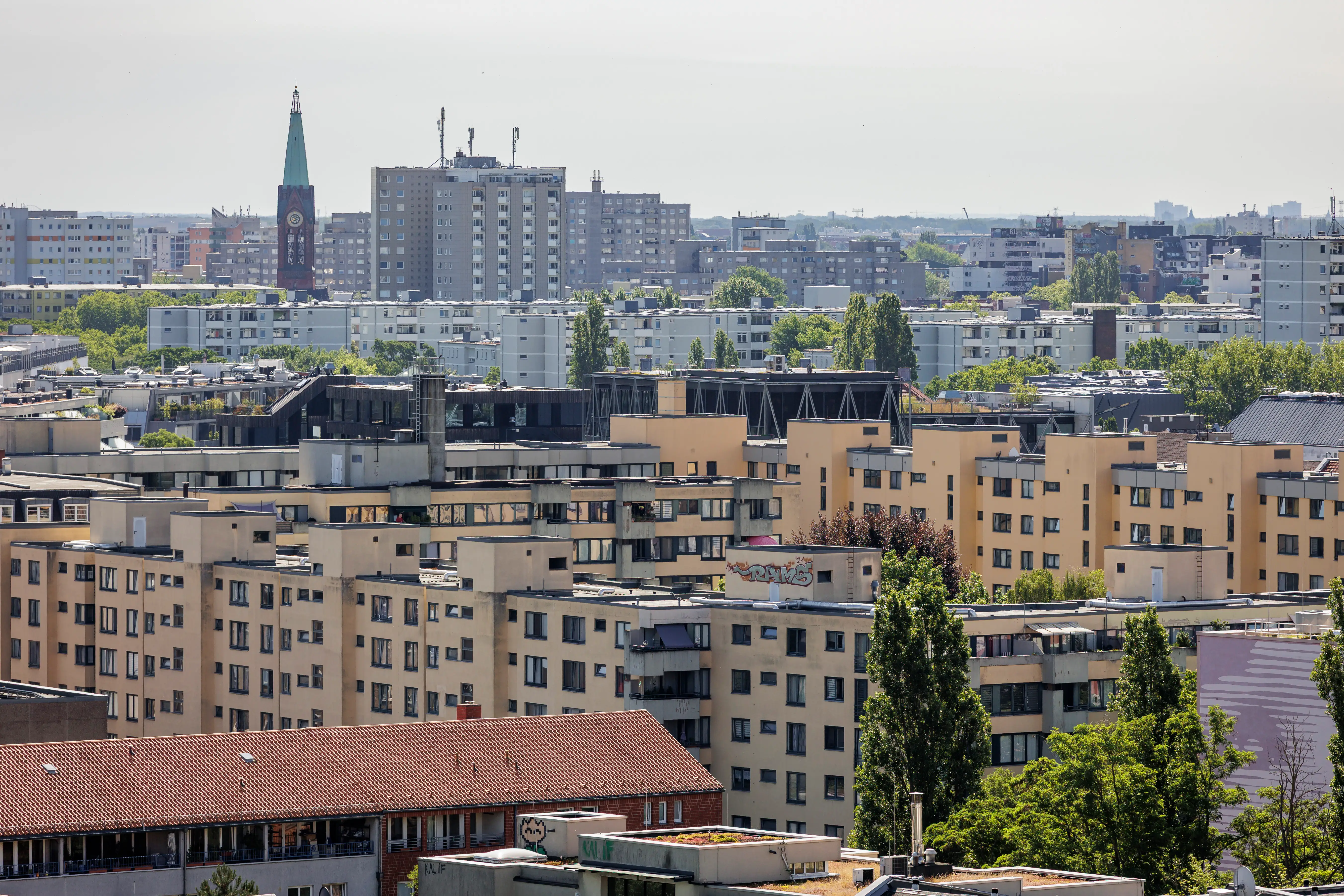 Dense urban apartment blocks and rooftops stretch toward a distant clock-tower church spire under a hazy sky.