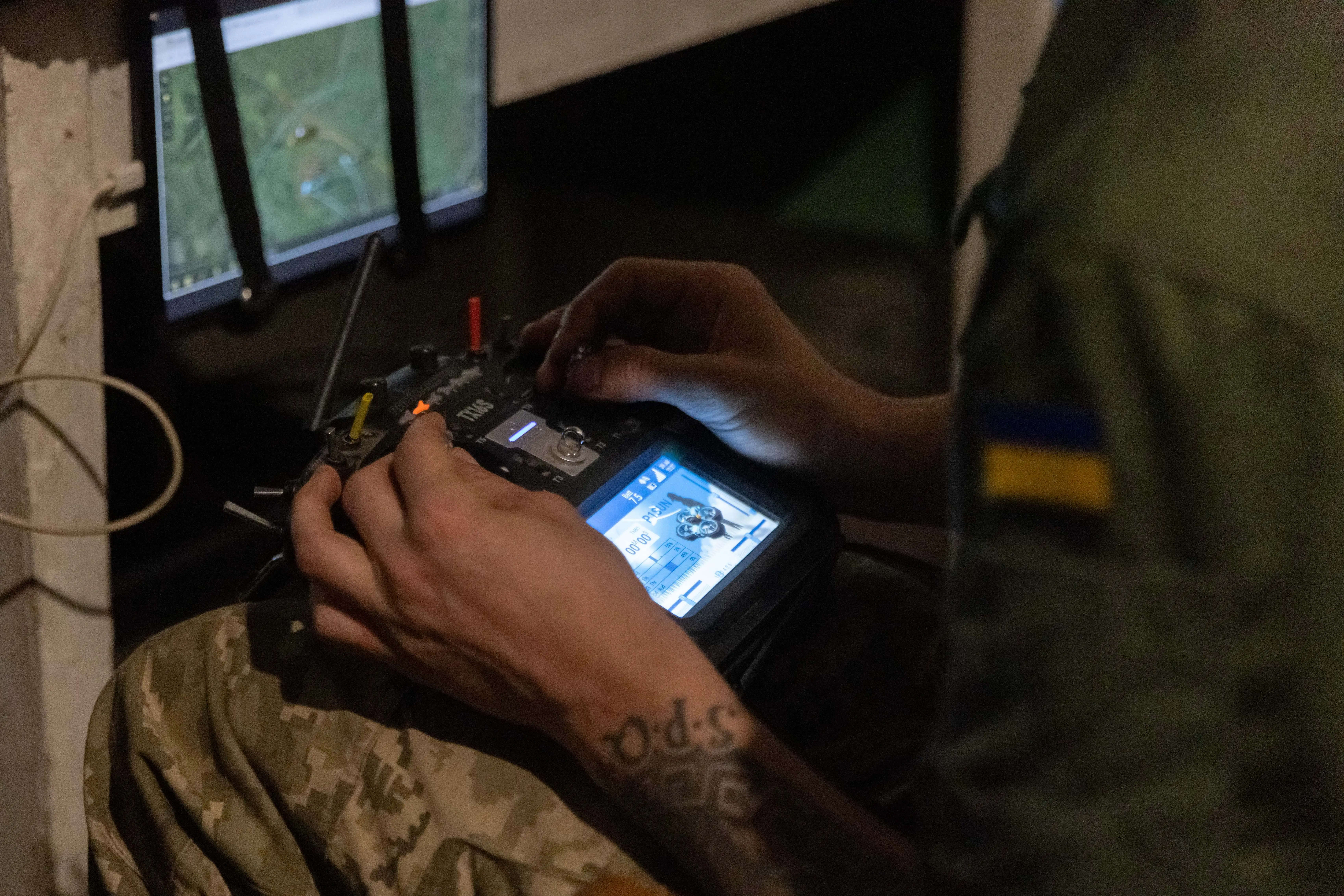 Uniformed person with a Ukrainian flag patch operates a drone controller beside monitors showing map and video feeds.