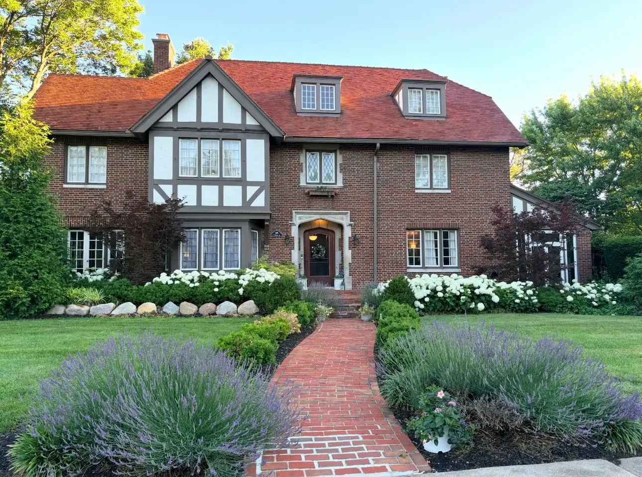 Large brick Tudor-style house with red tile roof, landscaped garden, and brick walkway leading to the front door.