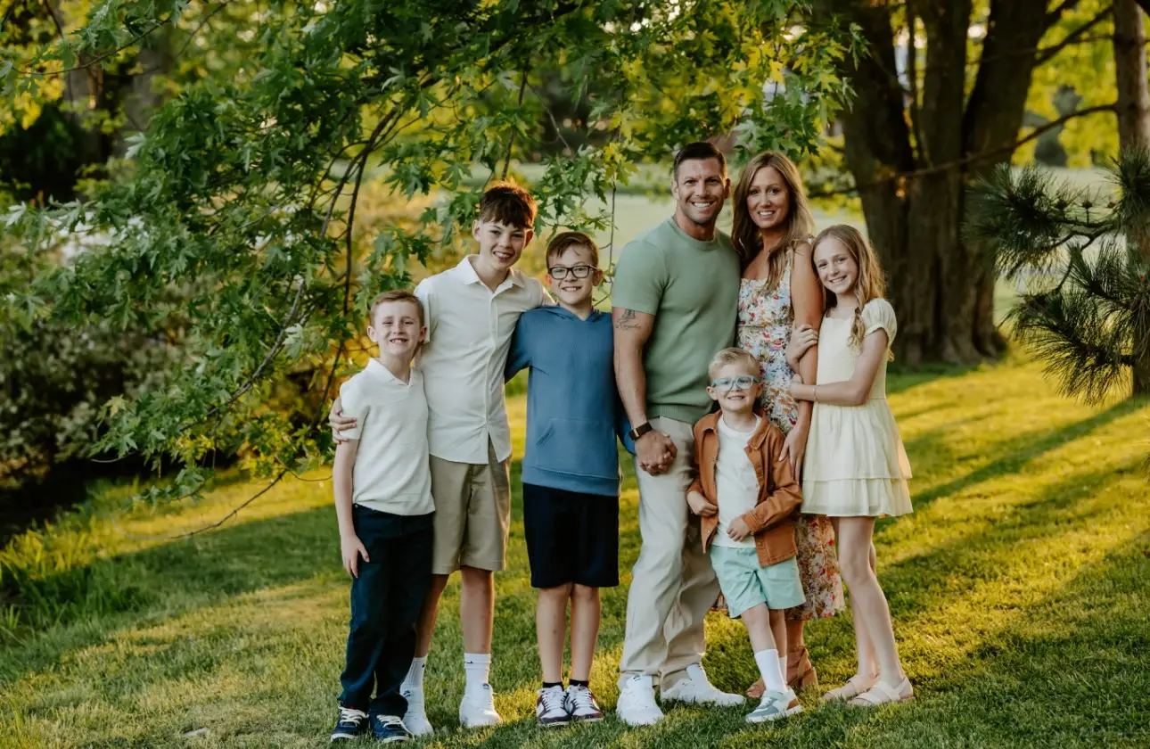 Seven people pose together on a sunlit grassy lawn beneath trees in an outdoor portrait.