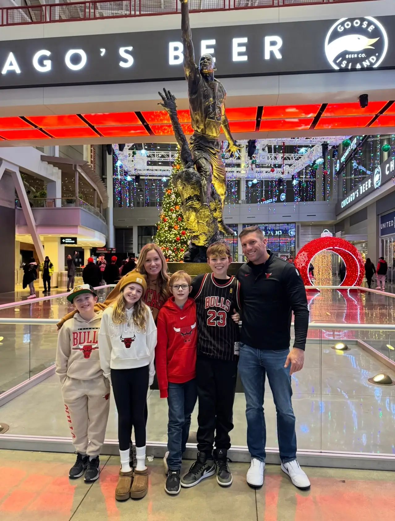 Six people in Chicago Bulls apparel pose before a basketball statue and holiday decorations inside an arena concourse.