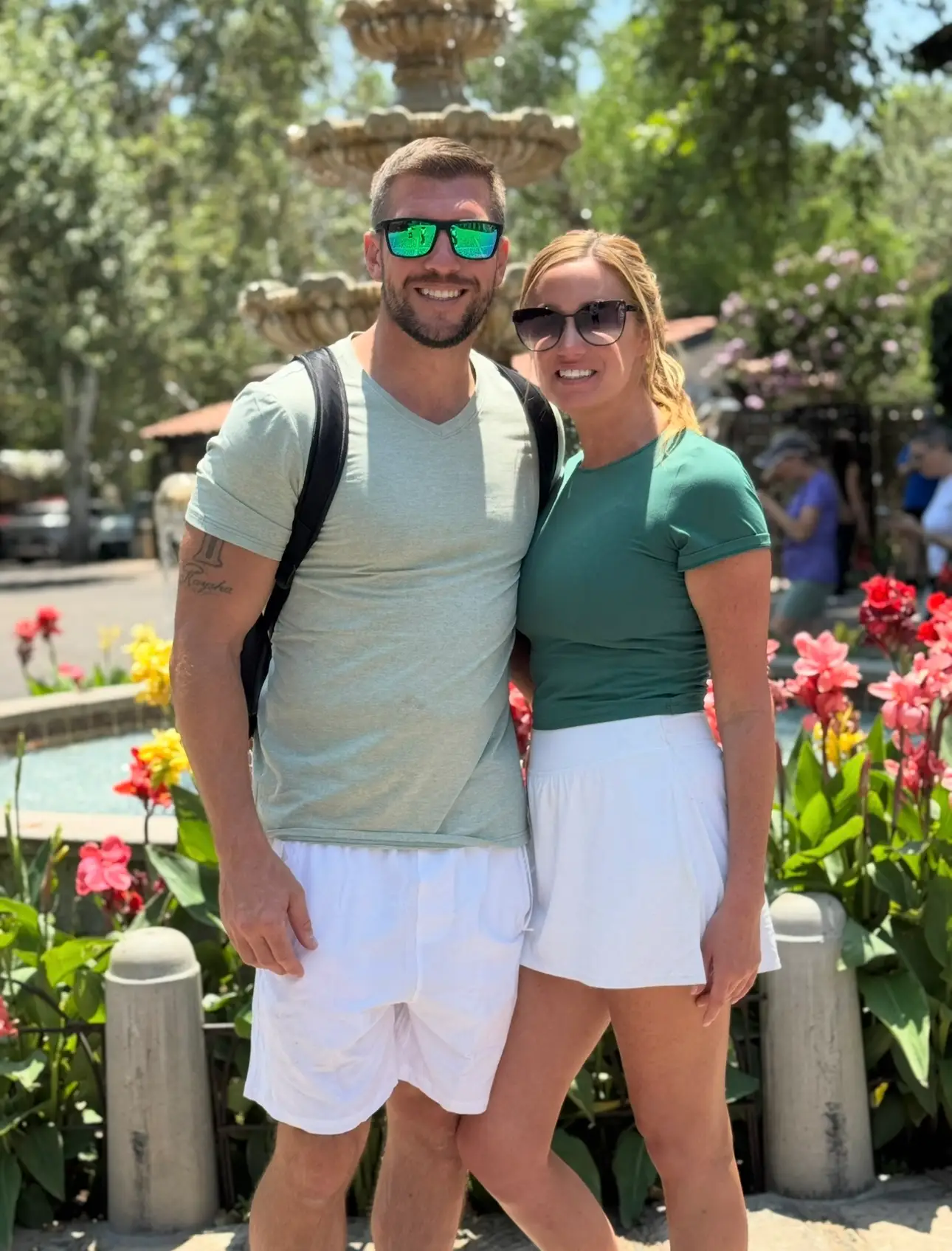 Two people wearing sunglasses pose together beside a fountain and colorful flowers in a sunny outdoor plaza.