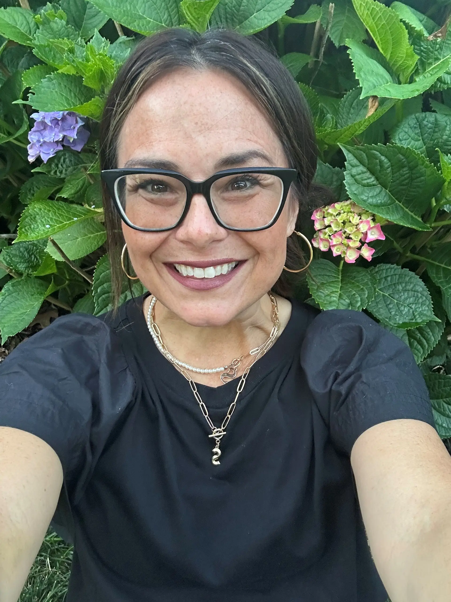 Smiling person wearing black glasses sits in front of green hydrangea leaves and purple flowers.