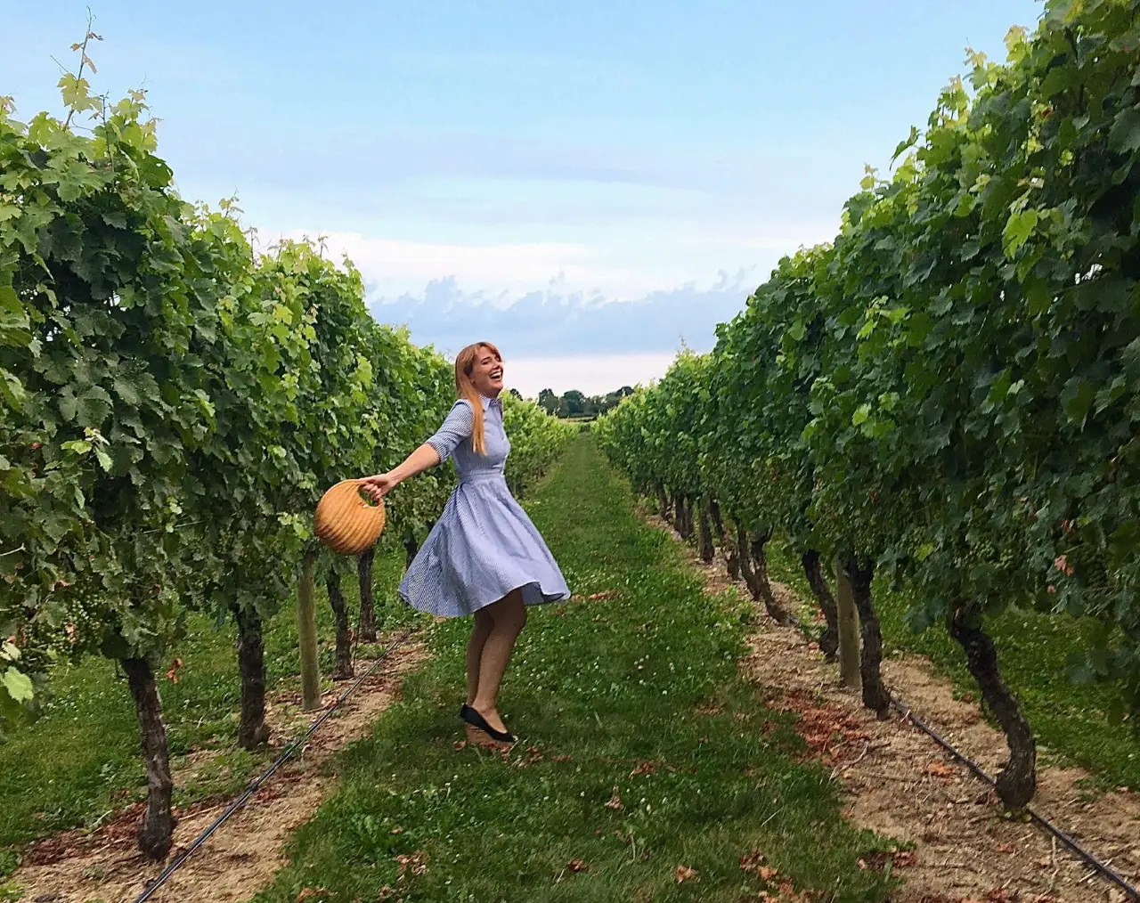 Woman smiling, twirling between rows of greenery under blue sky