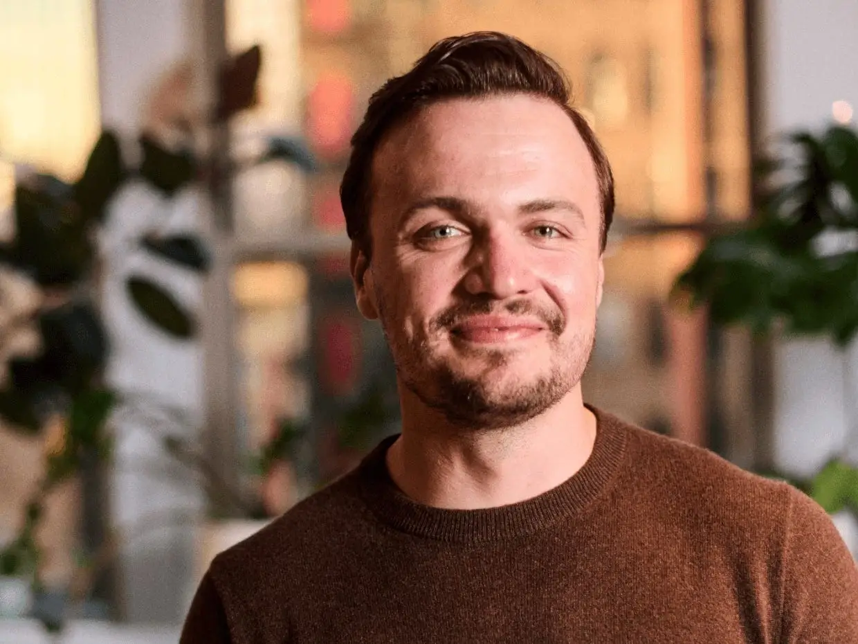 A smiling man in a brown sweater stands indoors near plants and large windows.